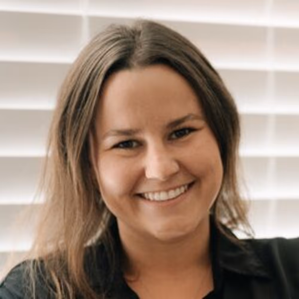 Smiling woman with long brown hair in front of white window blinds.