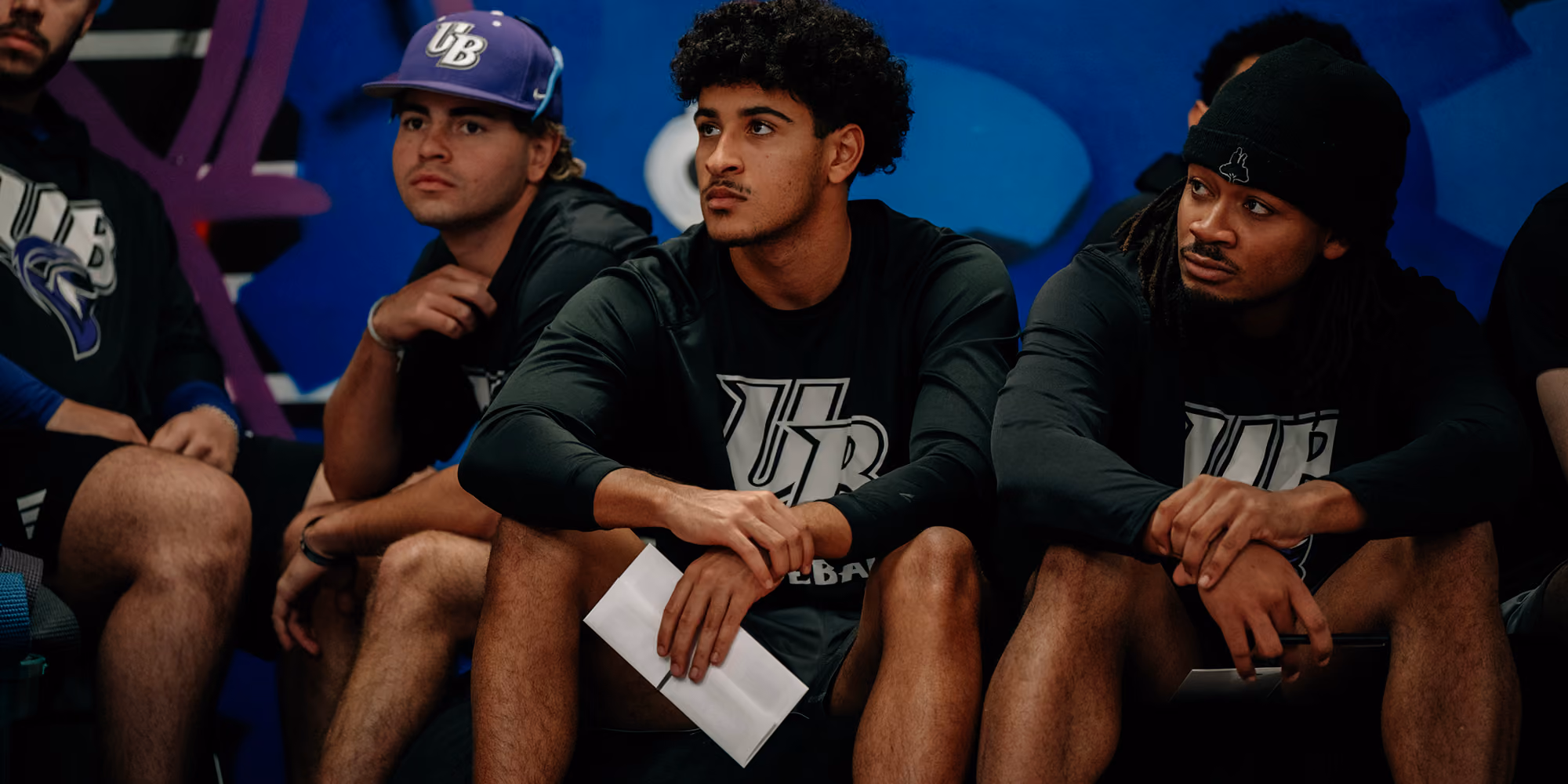 Three men in black UB shirts sitting and holding papers against a blue graffiti wall.