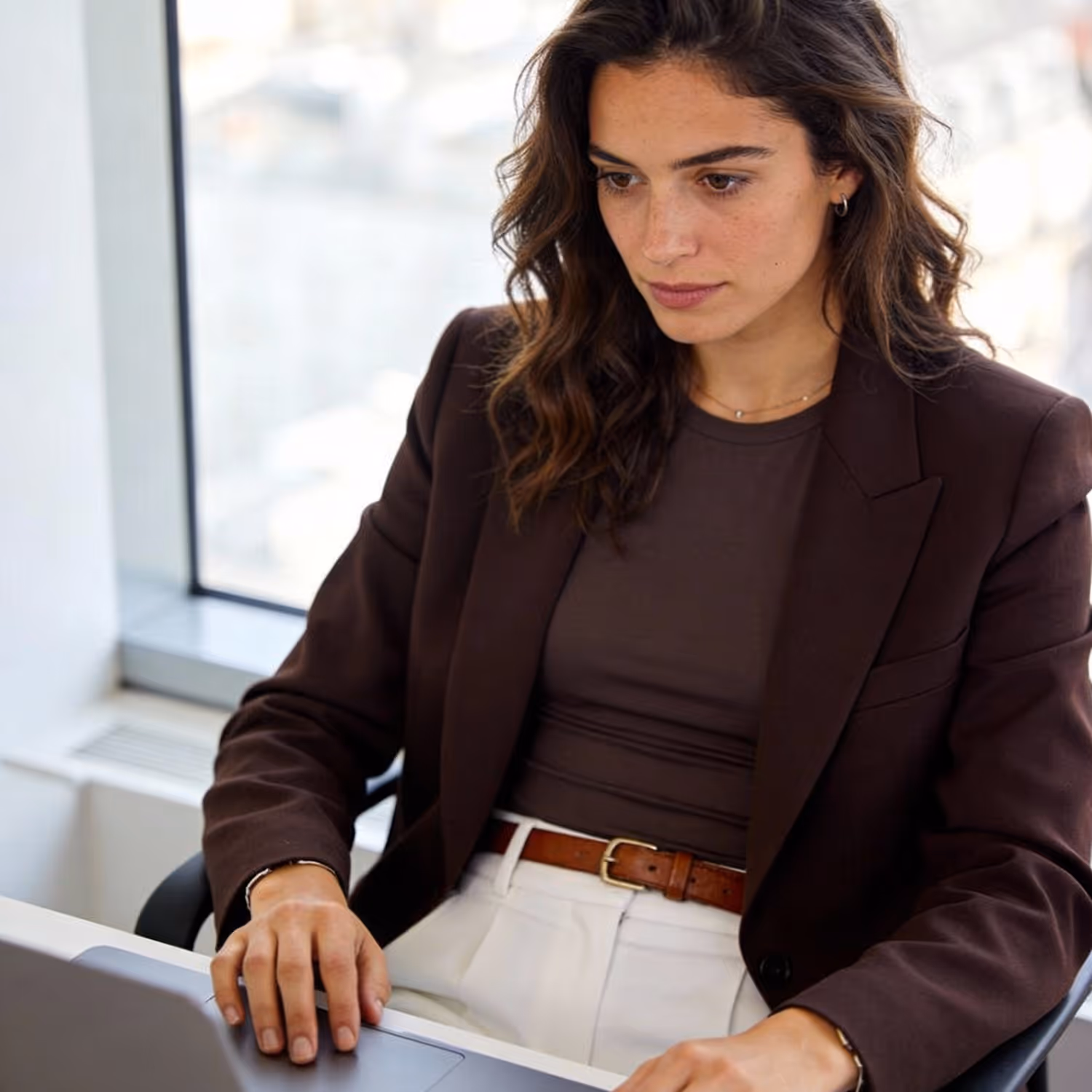 Woman with wavy hair in a brown blazer working on a laptop at a desk by a window.