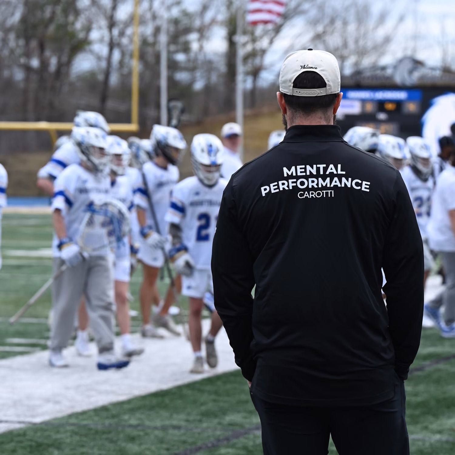 Coach wearing a hat and black jacket labeled Mental Performance Carotti facing lacrosse players on field.