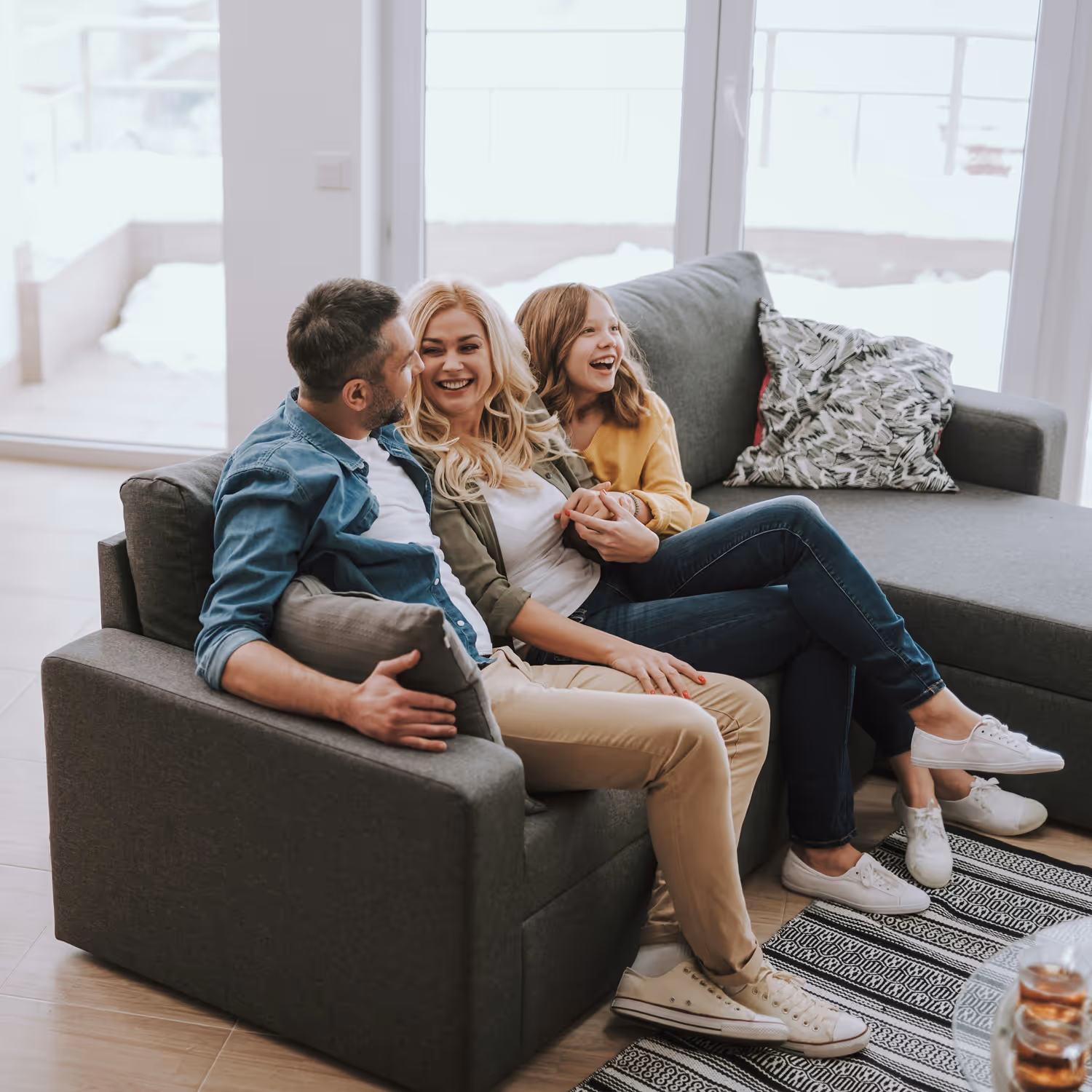 A smiling family of three sitting close on a gray sofa in a bright living room.