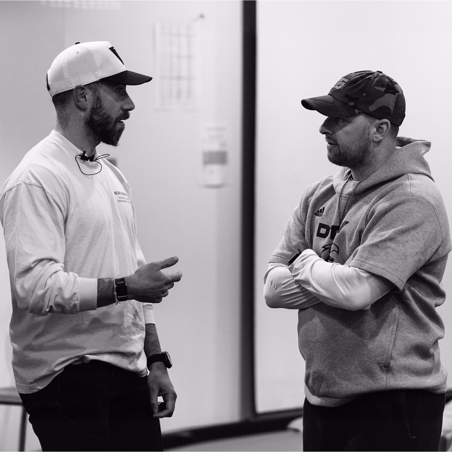 Two men wearing caps engaged in conversation indoors, one gesturing with his hand, the other with arms crossed.