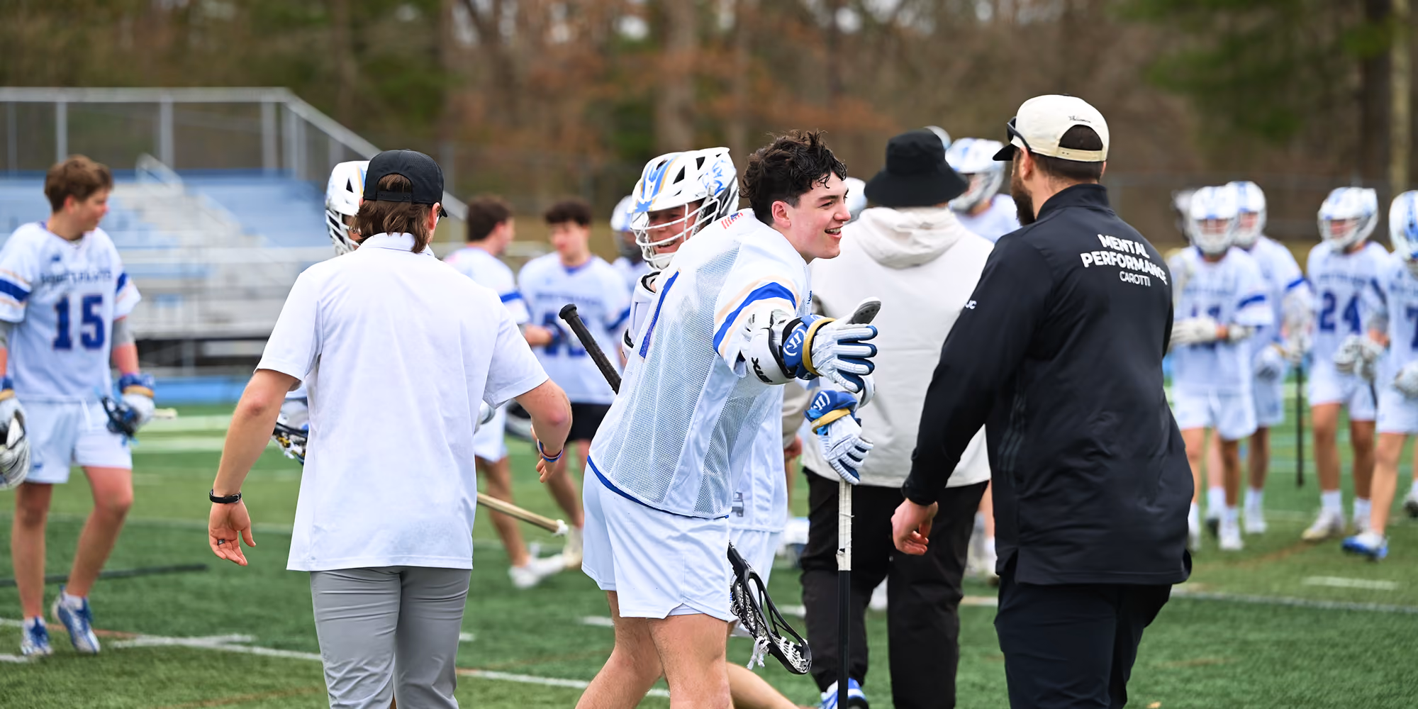 Lacrosse players in white and blue uniforms greet each other on a green field during a game.