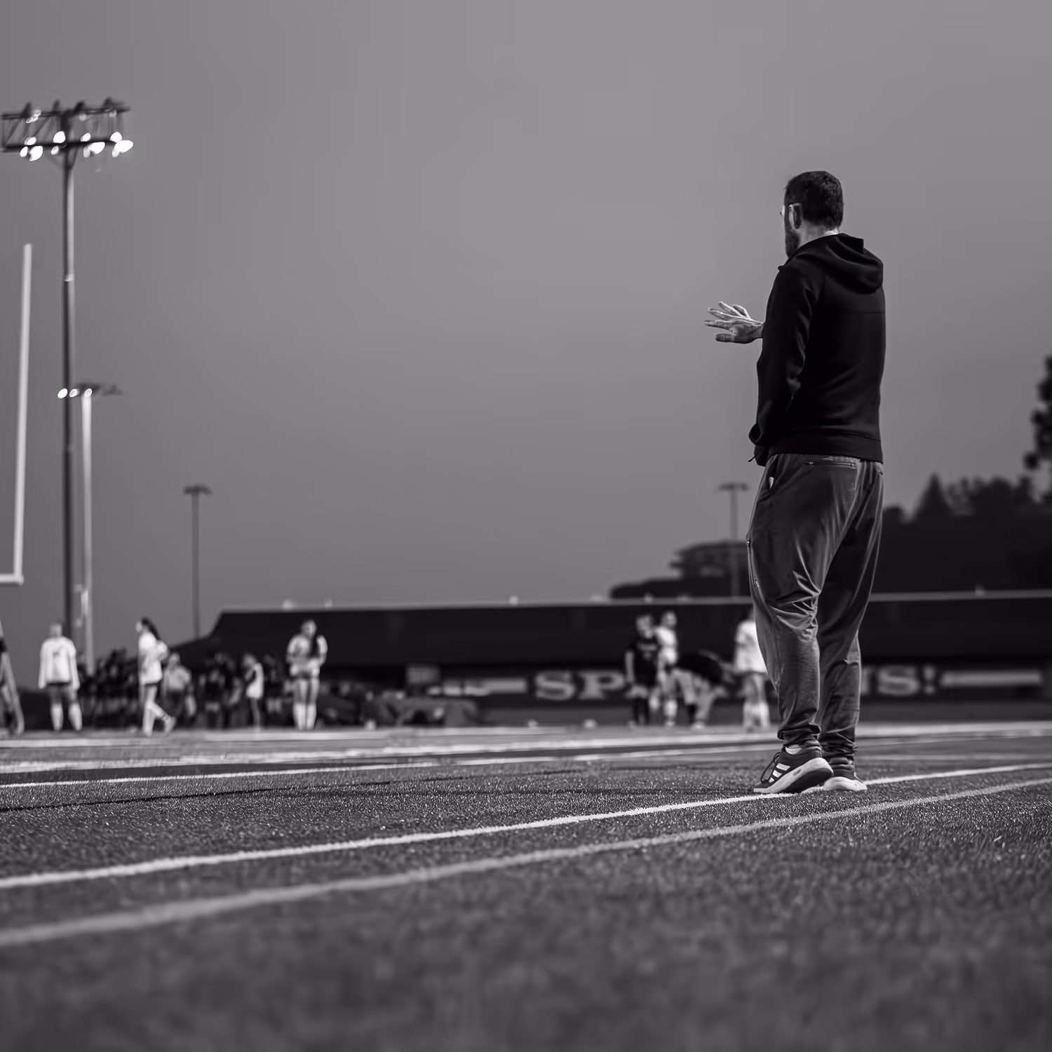 Coach on sideline clapping while players stand on a sports field in the background.