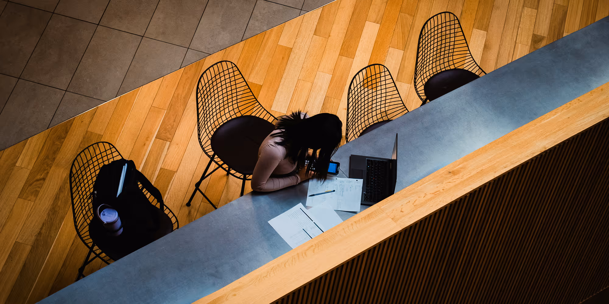 Person sitting at a counter with papers and laptop, using a smartphone, surrounded by wireframe chairs.