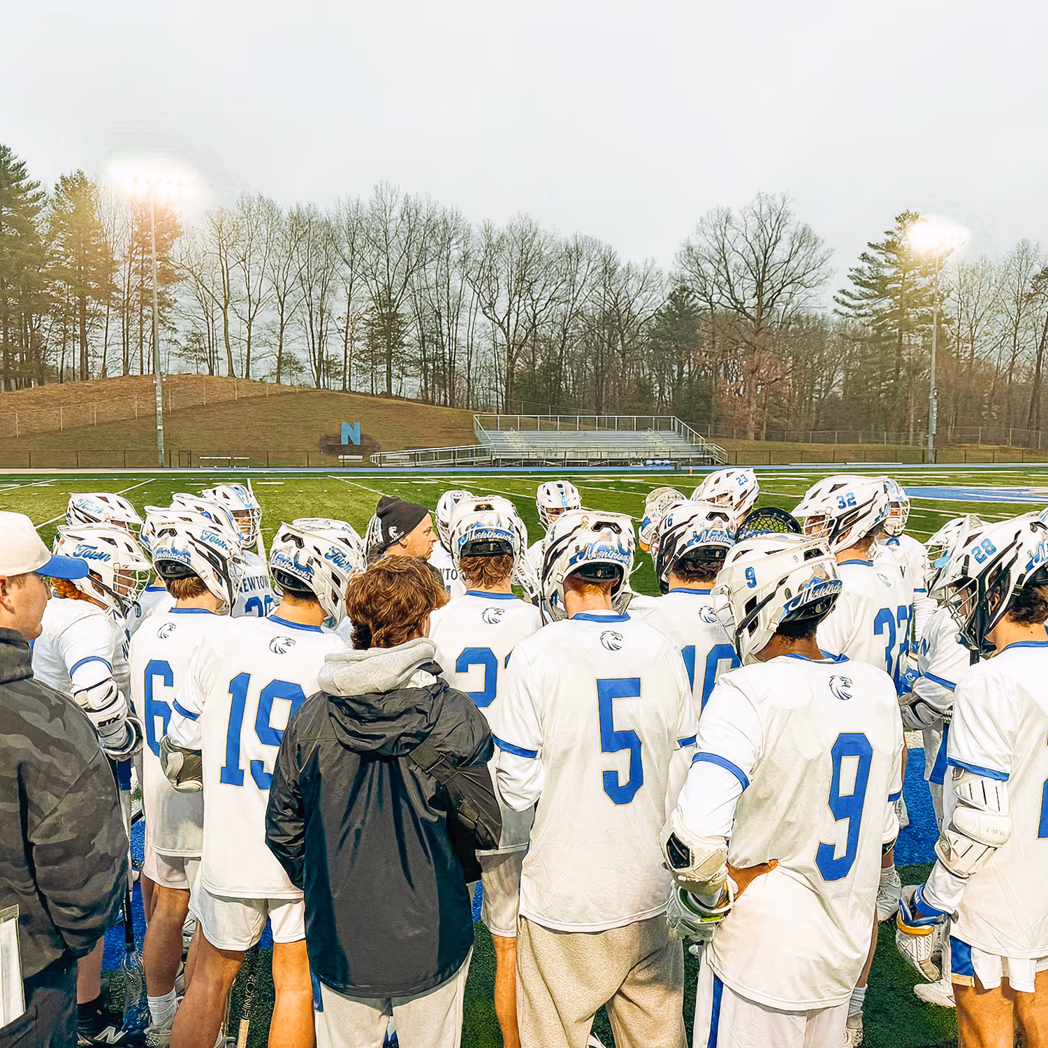 Lacrosse team in white uniforms and helmets gathered on a field with coaches during a game.