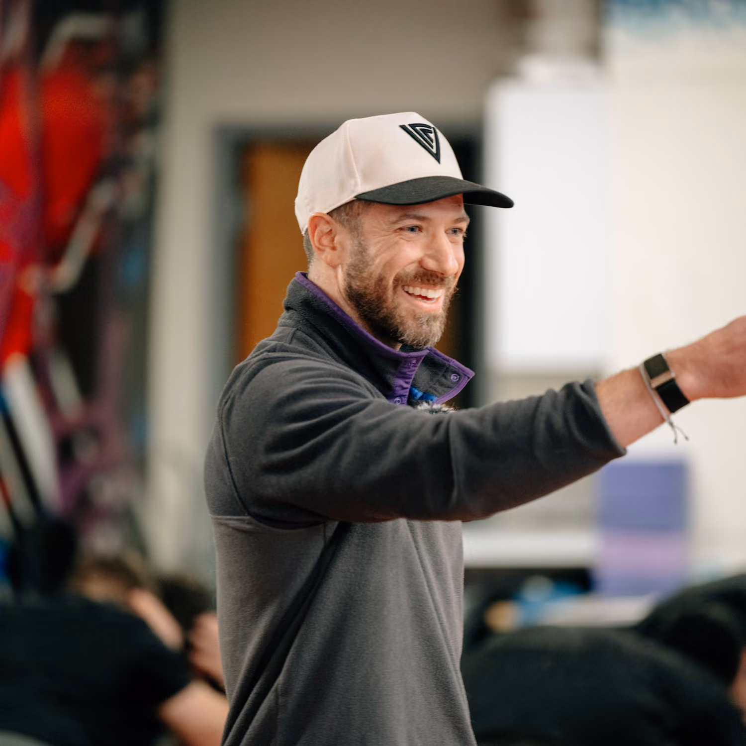 Smiling man with beard wearing a cap and dark jacket, reaching out with his arm in an indoor setting.