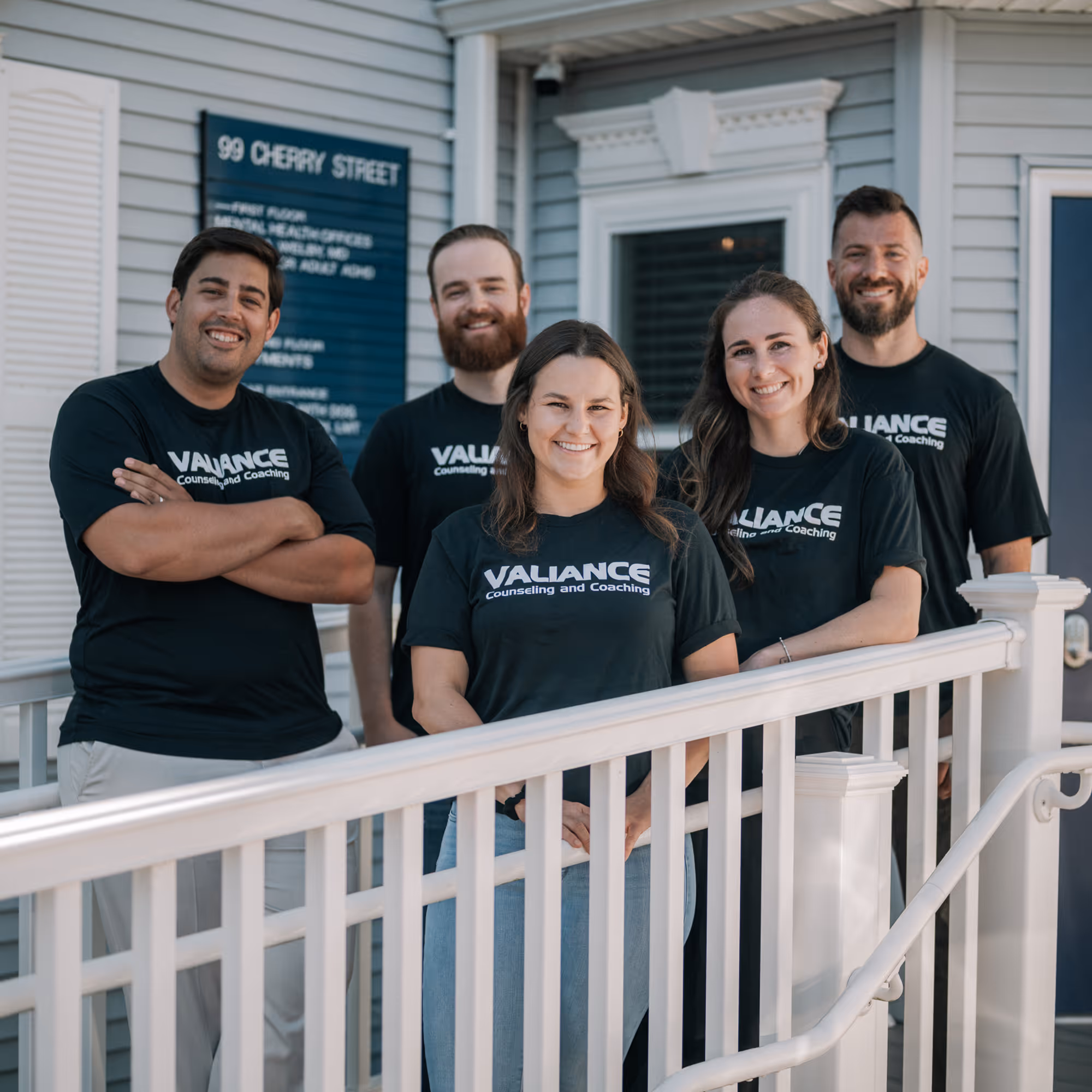 Five smiling people wearing black Valiance Counseling and Coaching shirts stand on a porch with white railing.