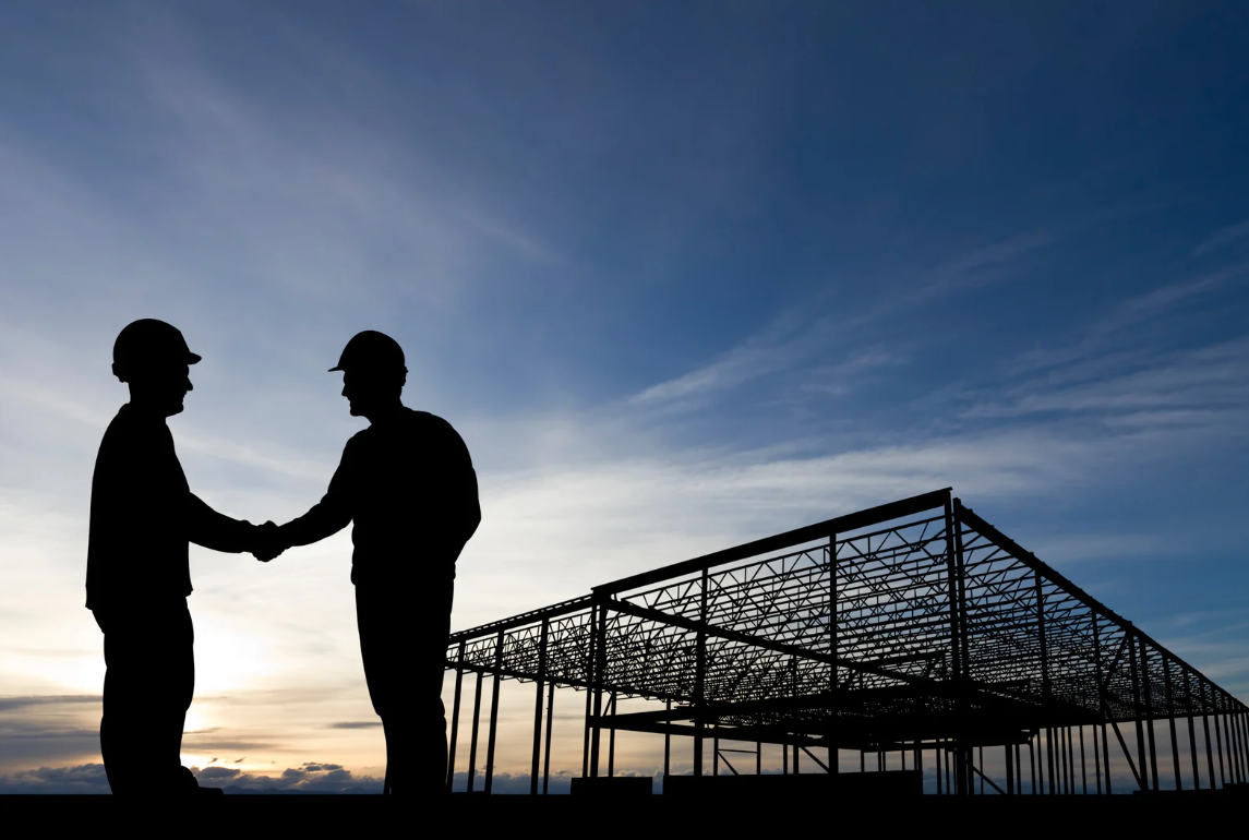 Zwei Bauarbeiter mit Schutzhelmen geben sich auf einer Baustelle bei Sonnenuntergang die Hand.