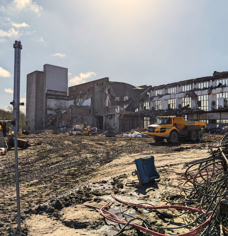 Baustelle mit teilweise abgerissenem Industriegebäude, Baufahrzeug und Baustellenausrüstung bei Sonnenschein.