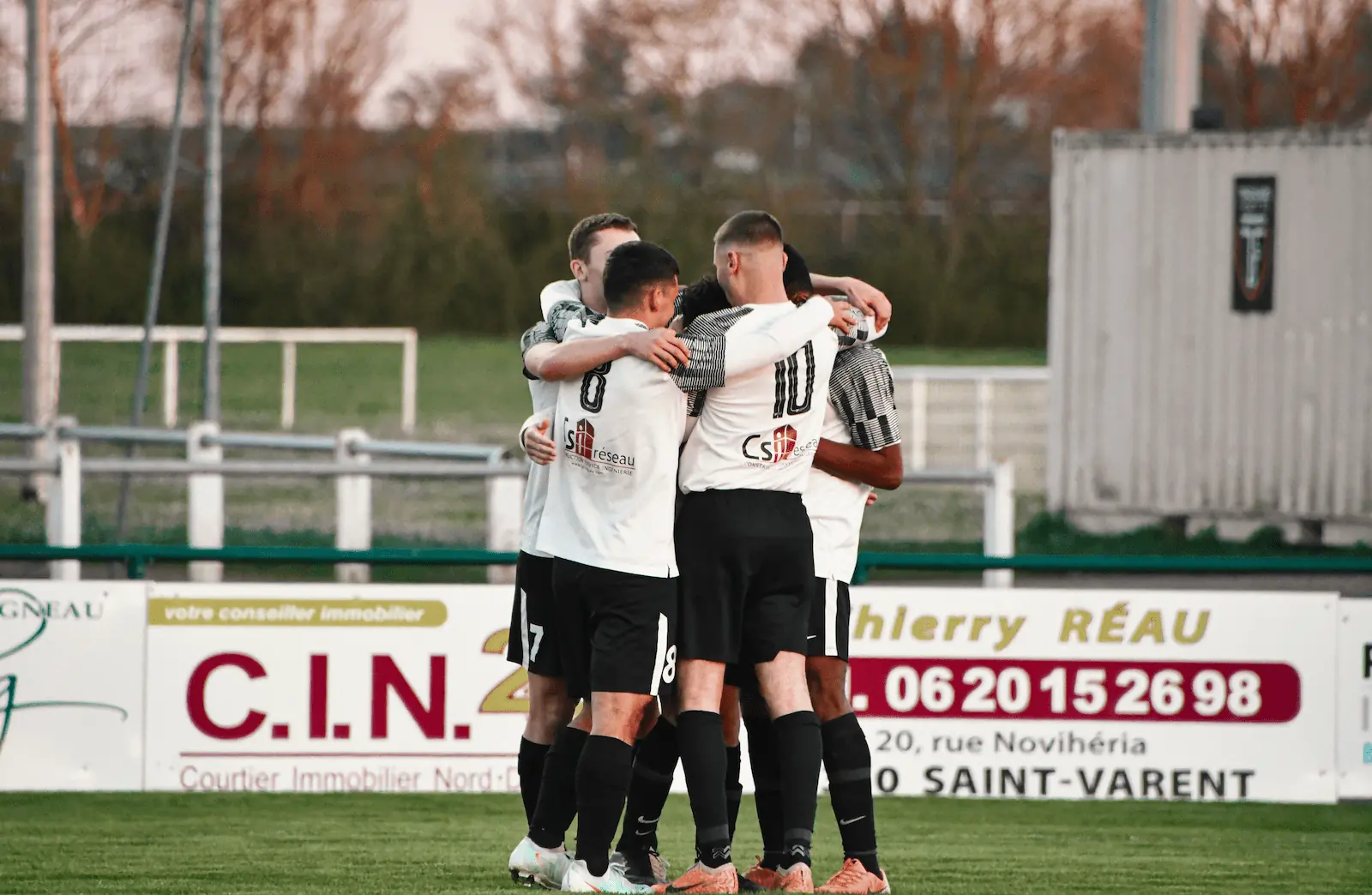Photographie d’une équipe de football masculine de la Ligugéenne se regroupant en cercle sur le terrain, sur leur maillot on aperçoit leur sponsor, CSi réseau. 