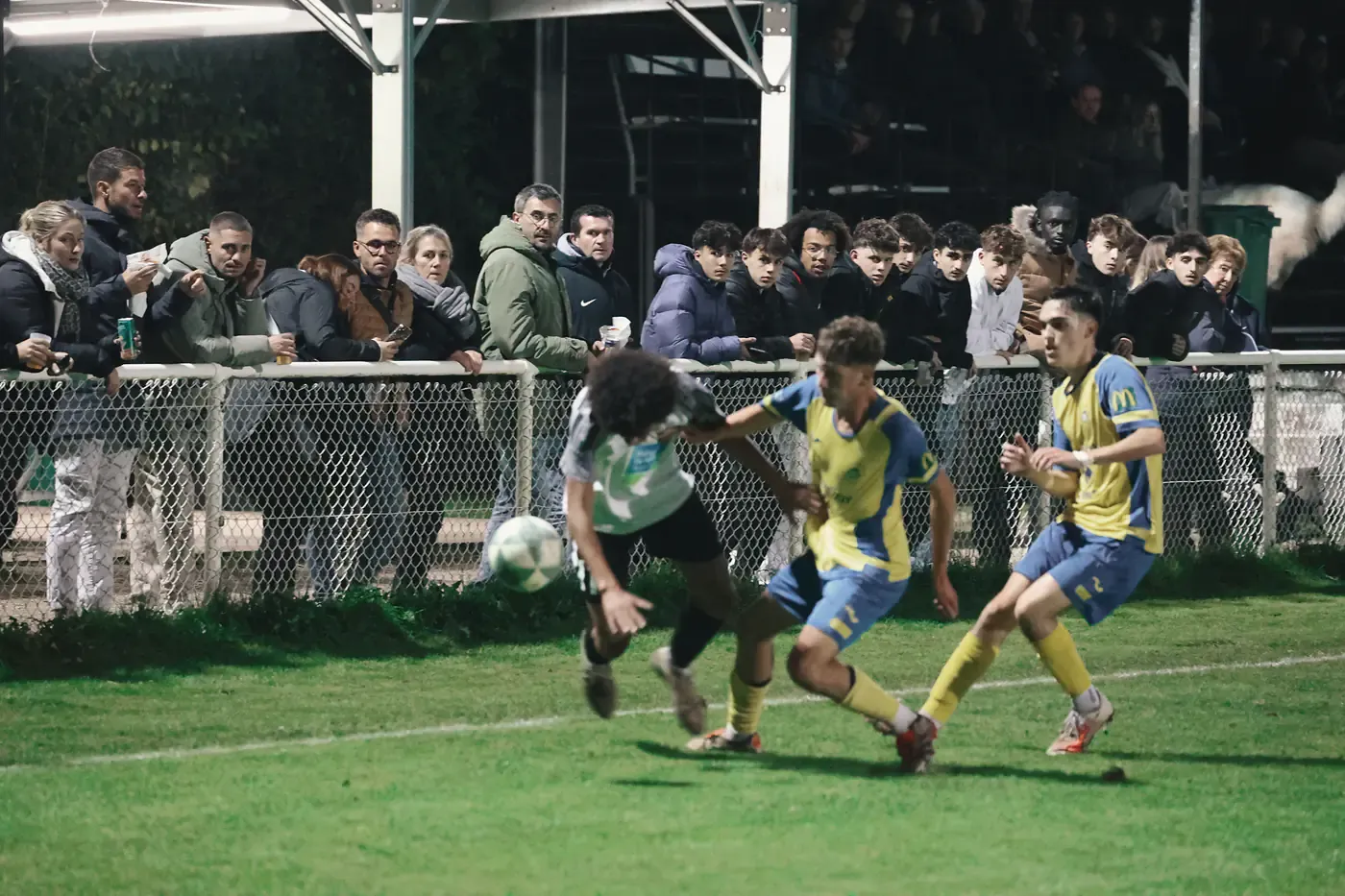 Photo d'une action durant un match de football au premier plan. Les supporters observent l'action au second plan.