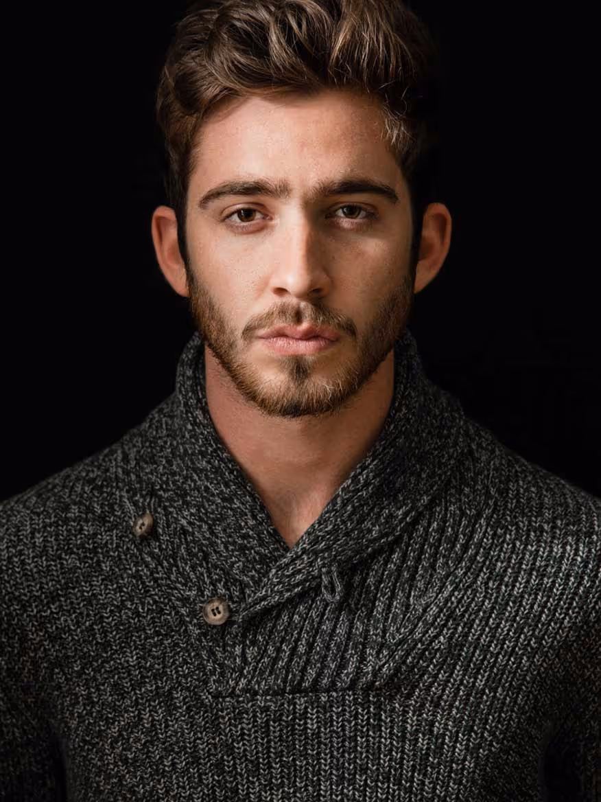 Portrait of a young man with short brown hair and a beard wearing a dark gray knitted sweater with buttons on the collar against a black background.
