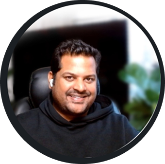 Smiling man with short dark hair wearing a black hoodie and wireless earbuds sitting in a chair with a blurred indoor background.