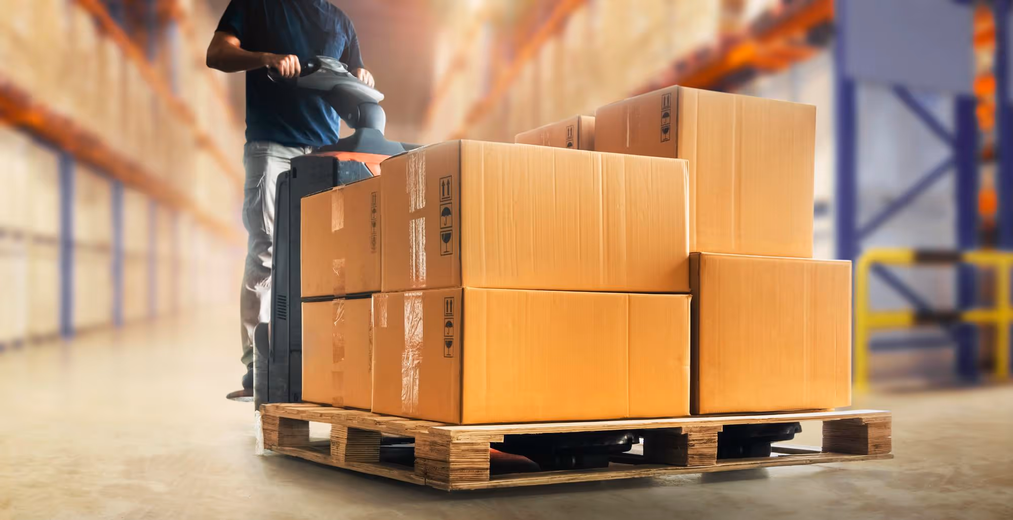 Man operating pallet jack moving stacked cardboard boxes on a wooden pallet in a warehouse.