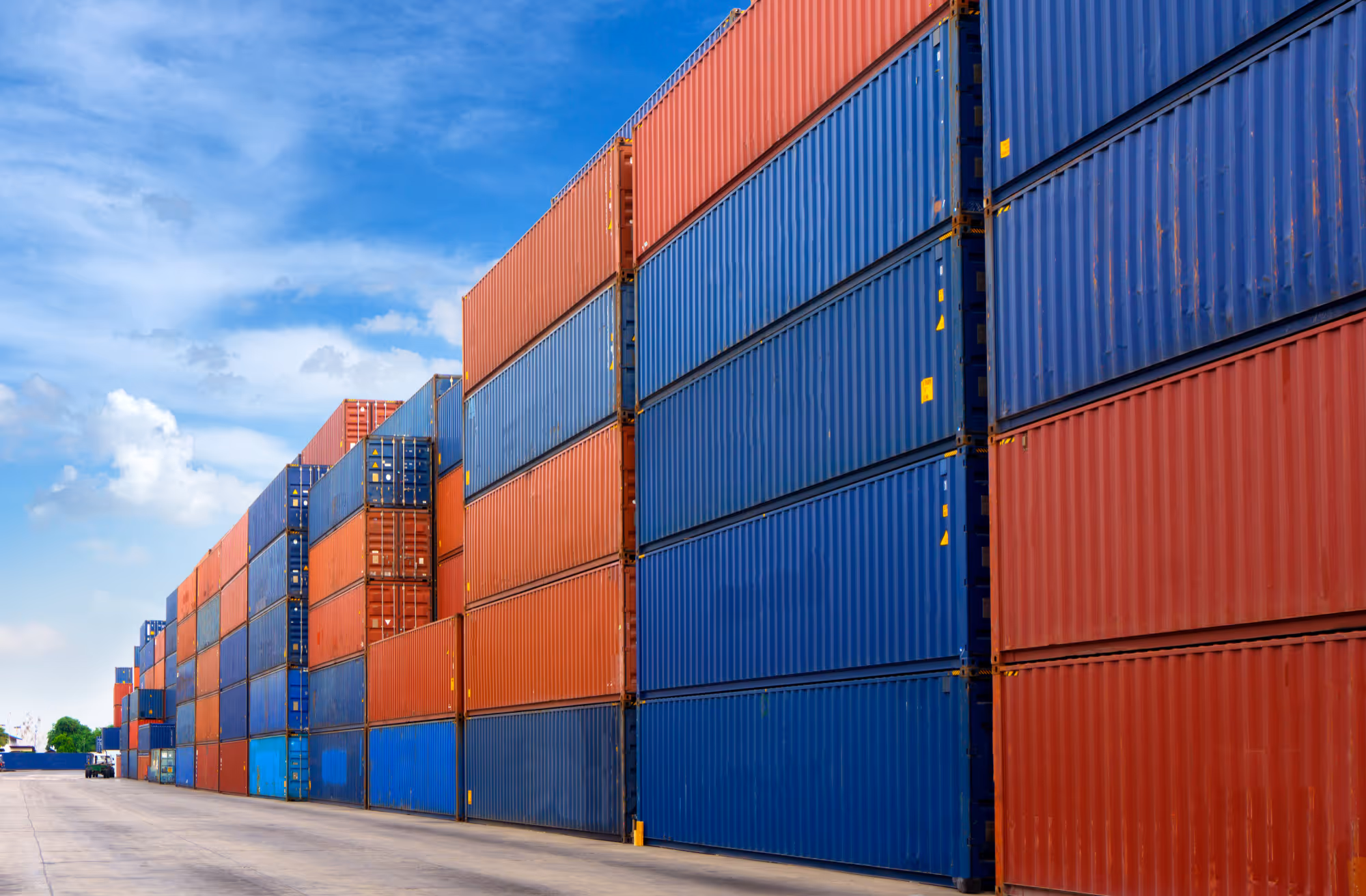 Rows of stacked red and blue shipping containers under a partly cloudy blue sky.