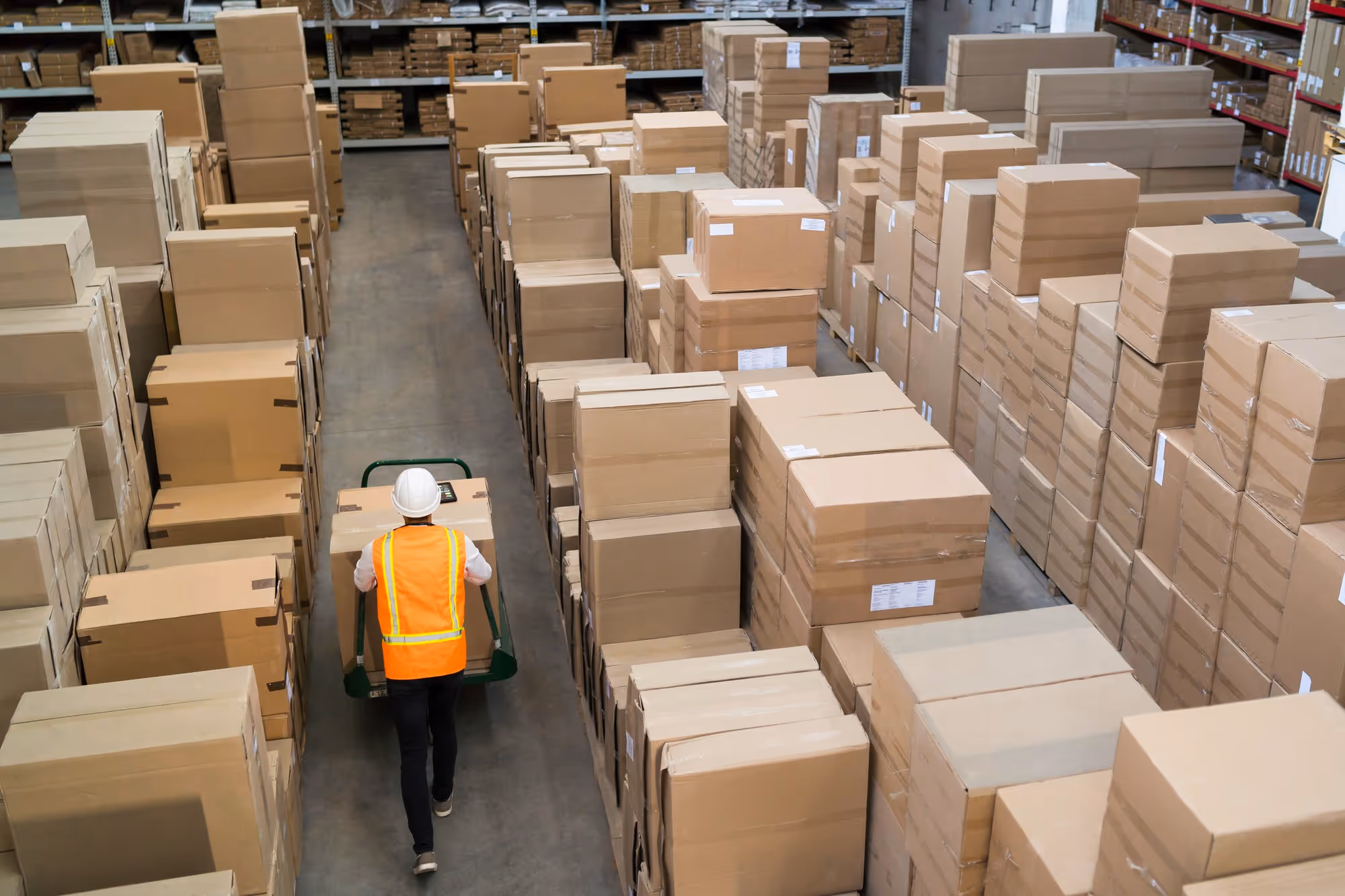 Warehouse worker in safety vest and helmet pushing a cart with boxes through aisles of stacked cardboard boxes.