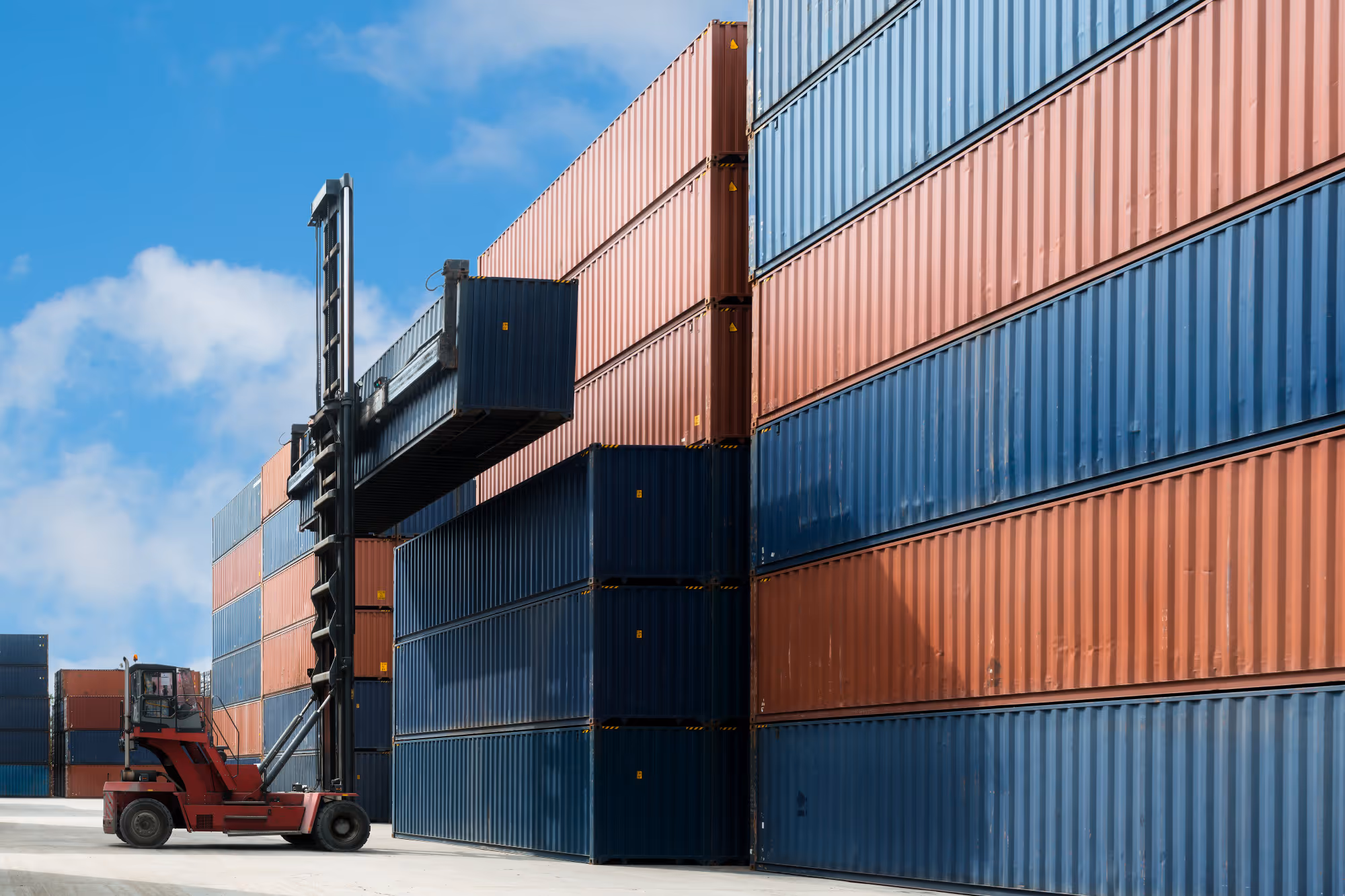 Red forklift lifting a blue shipping container among stacked blue and orange containers under a blue sky.