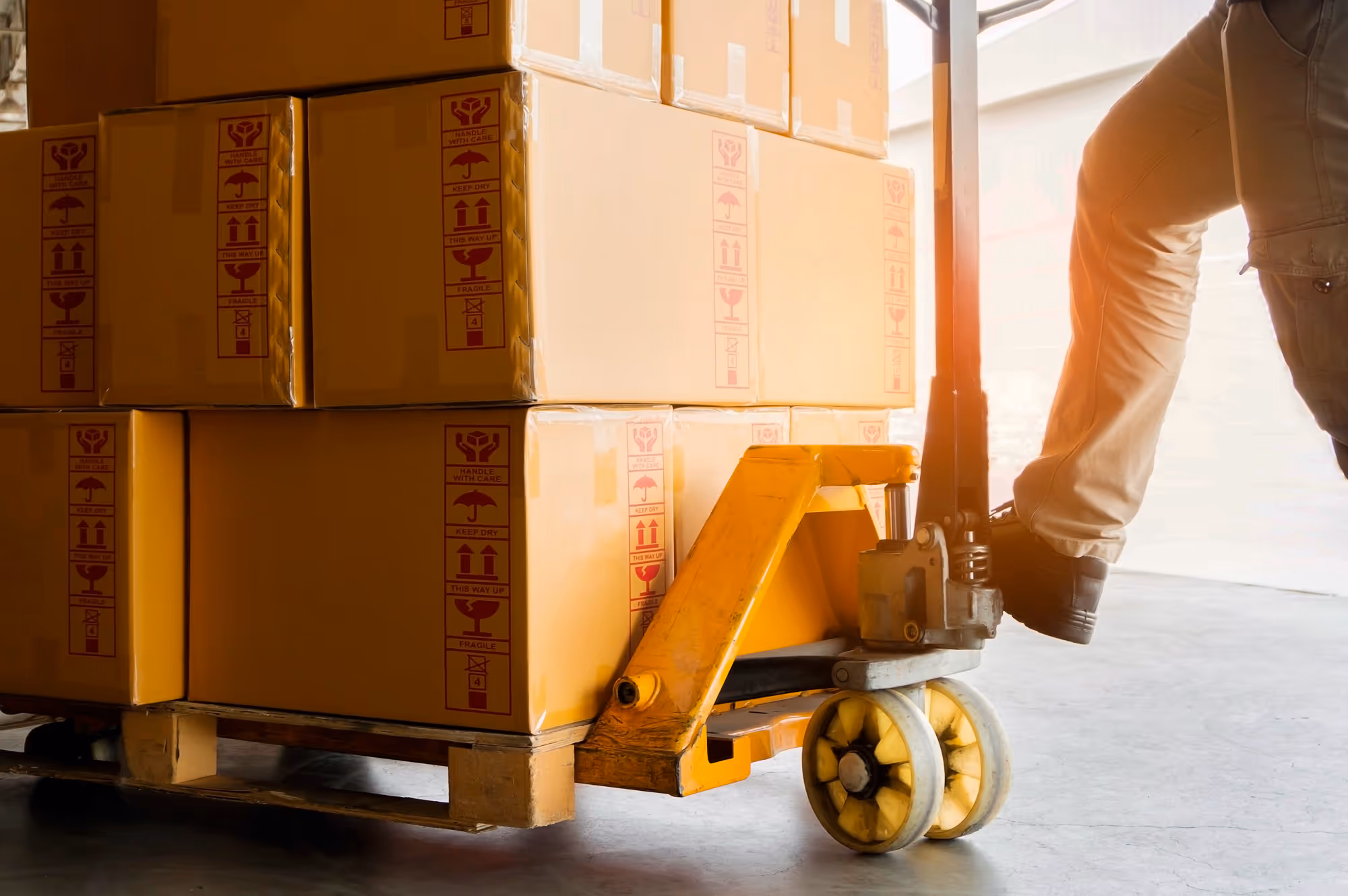 Person using a yellow pallet jack to move a wooden pallet stacked with cardboard boxes.