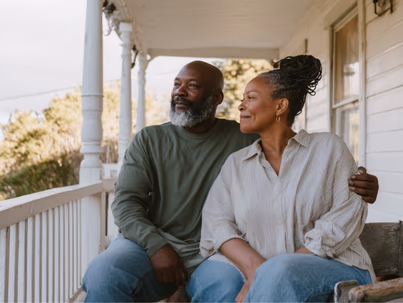 Middle-aged couple sitting closely on a porch bench, looking content and gazing into the distance.