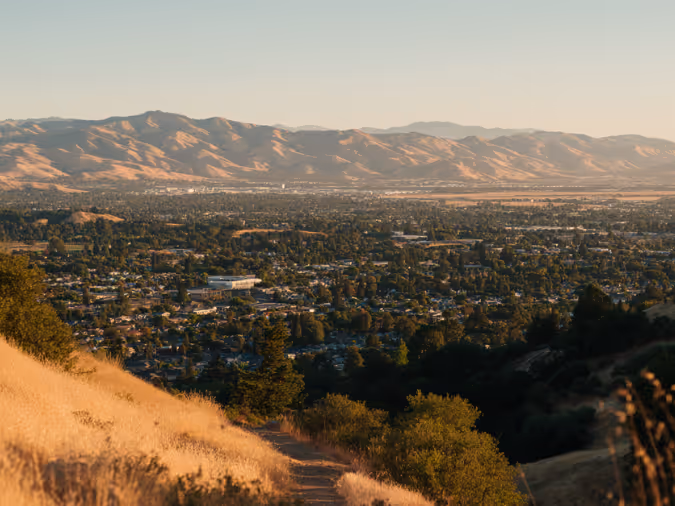View overlooking a city with a dry golden grassy hill in the foreground and mountain range in the background under a clear sky.