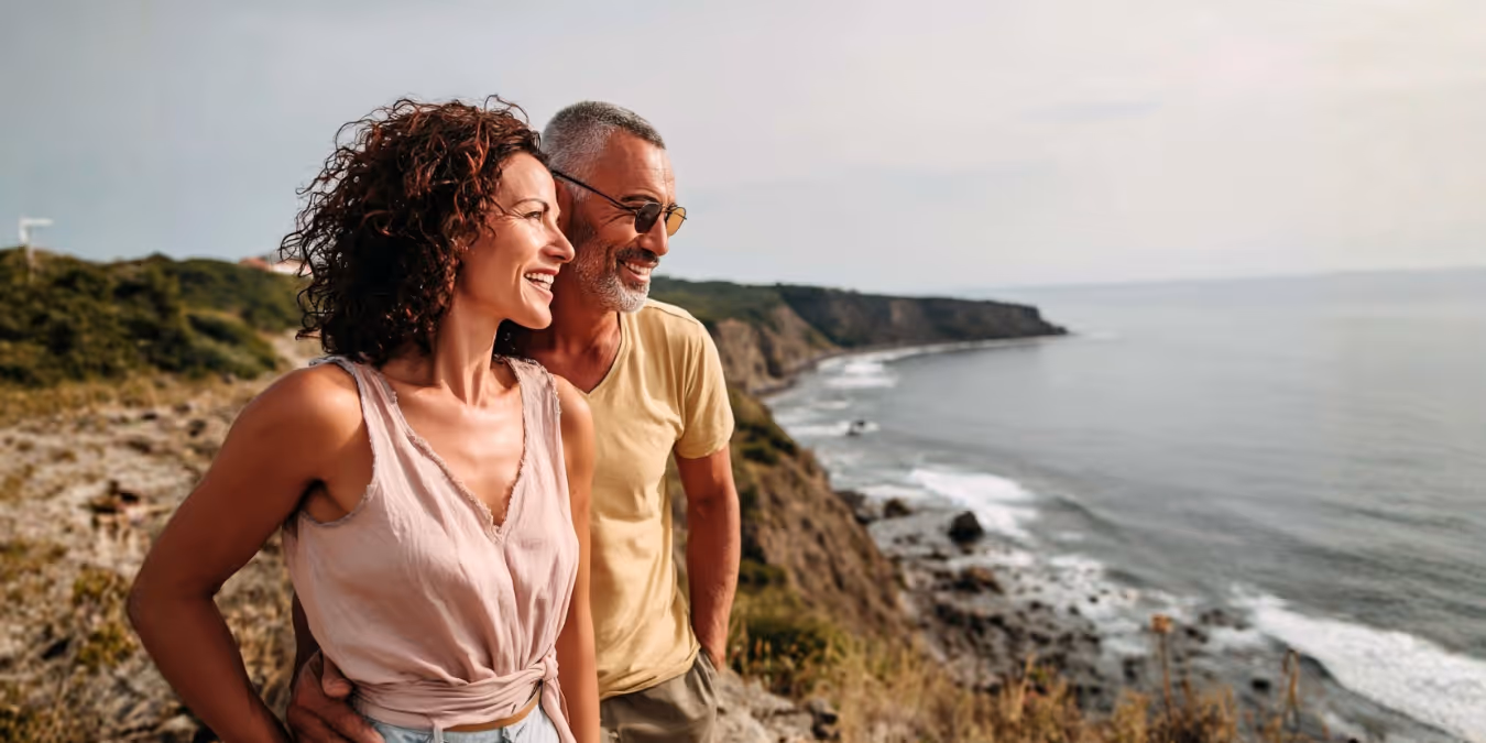 Smiling couple standing on a rocky cliff overlooking the ocean and coastline.