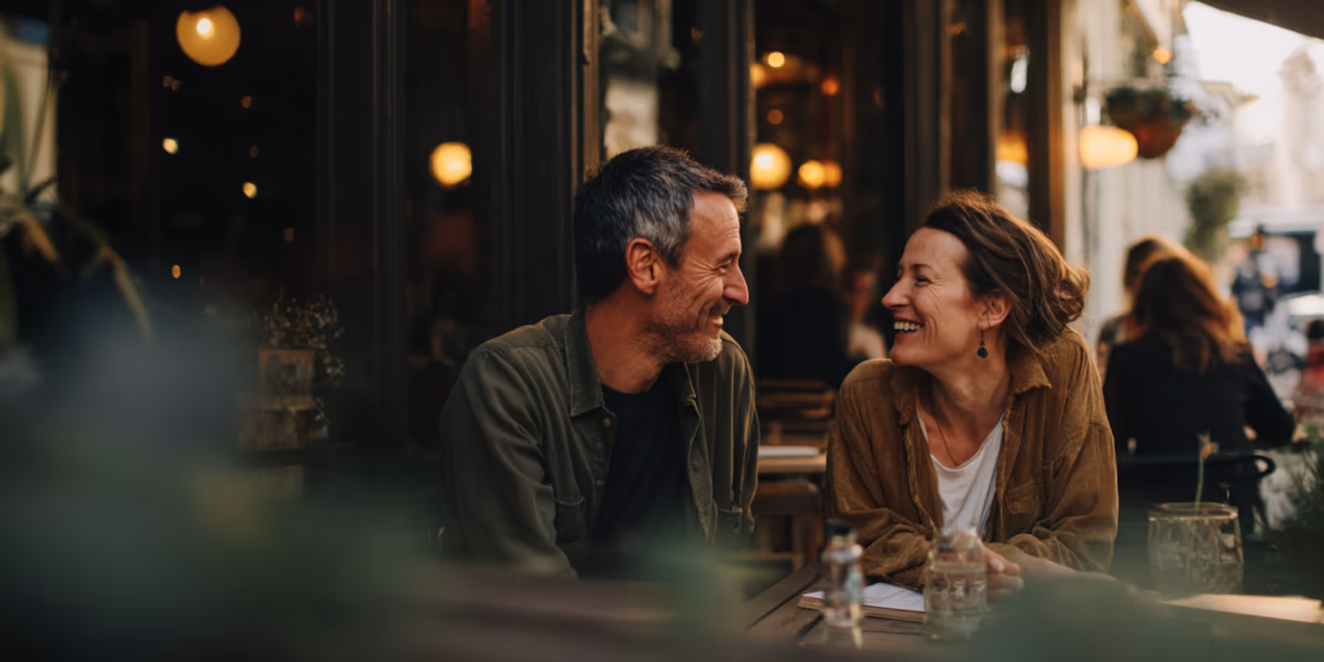 Middle-aged couple smiling and looking at each other while sitting at a table in a warmly lit outdoor cafe.