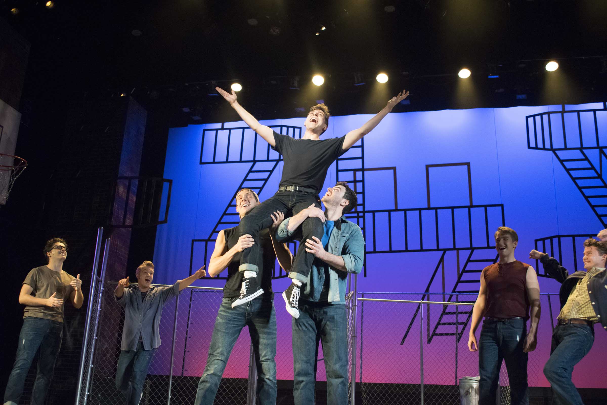 Theatrical scene with seven male actors, one raised by two others, against a set with fence and fire escapes.