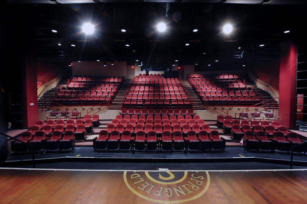 Empty theater with red seats facing a stage with ceiling spotlights on.