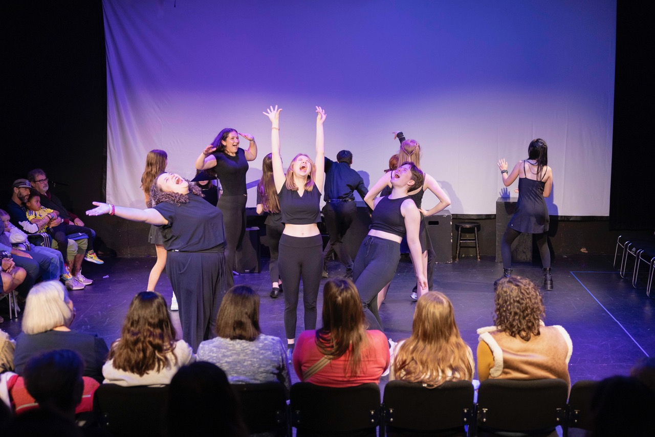 Group of performers on stage in black outfits with audience watching in front rows.