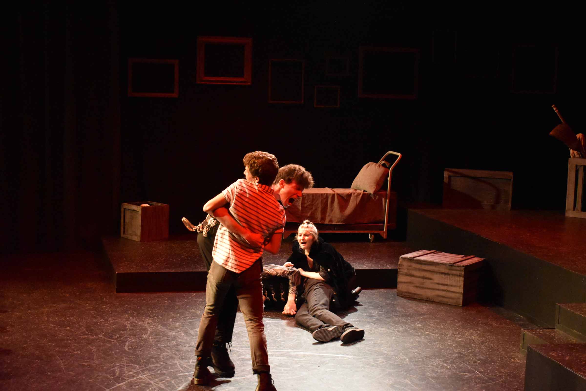 Two people hugging on stage while a woman kneels beside a person lying on the floor in a dramatic scene.