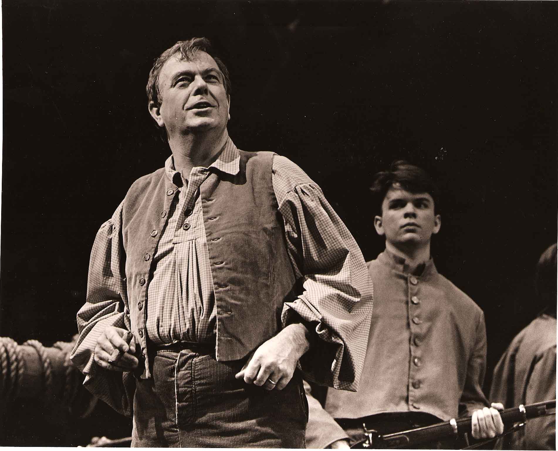 Man in period costume speaking with young man holding a rifle behind him on stage.