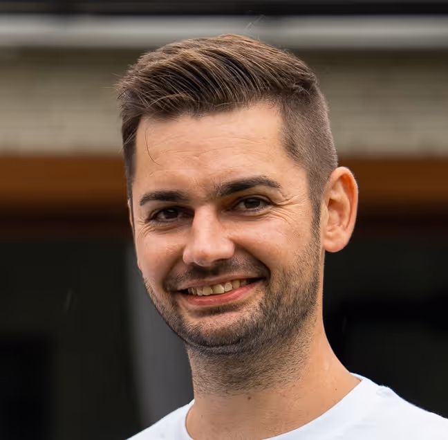 Mann mit kurzem braunem Haar und Bart lächelt, trägt weißes T-Shirt vor unscharfem Hintergrund.