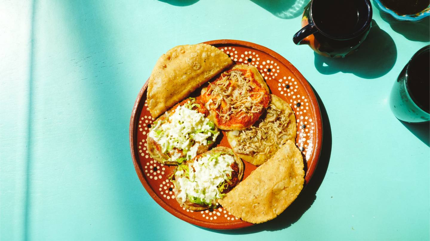 Traditional Mexican plate with three sopes topped with cheese, lettuce, and shredded meat, accompanied by two folded empanadas on a turquoise table.