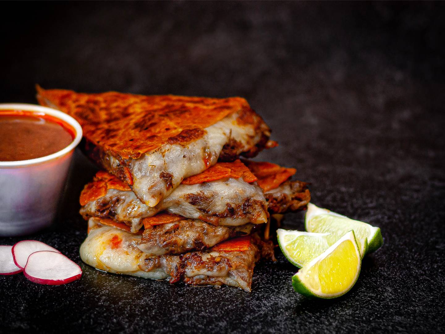 Stack of cheesy, crispy quesadilla slices with a cup of red sauce, lime wedges, and radish slices on a dark surface.