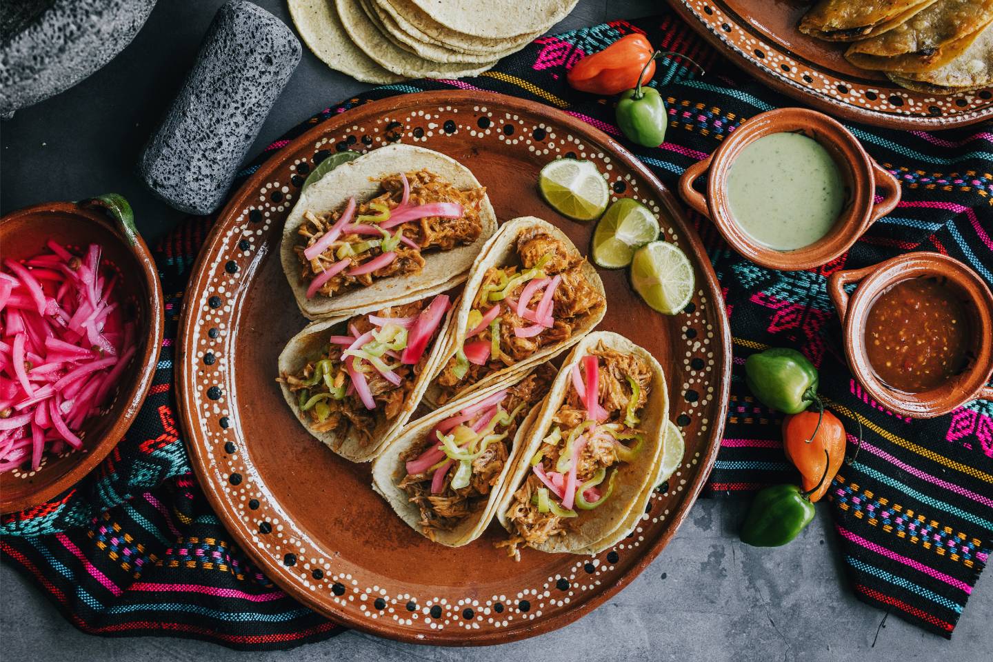 Four tacos filled with shredded meat and pickled vegetables on a decorated clay plate with lime wedges, surrounded by sauces, fresh tortillas, and chili peppers on a colorful cloth.