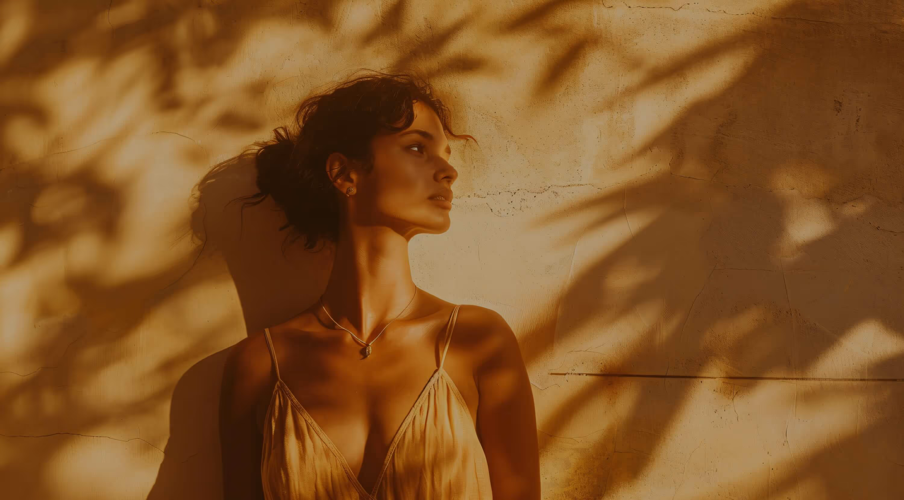Woman in a beige dress standing against a textured wall with shadows of leaves.