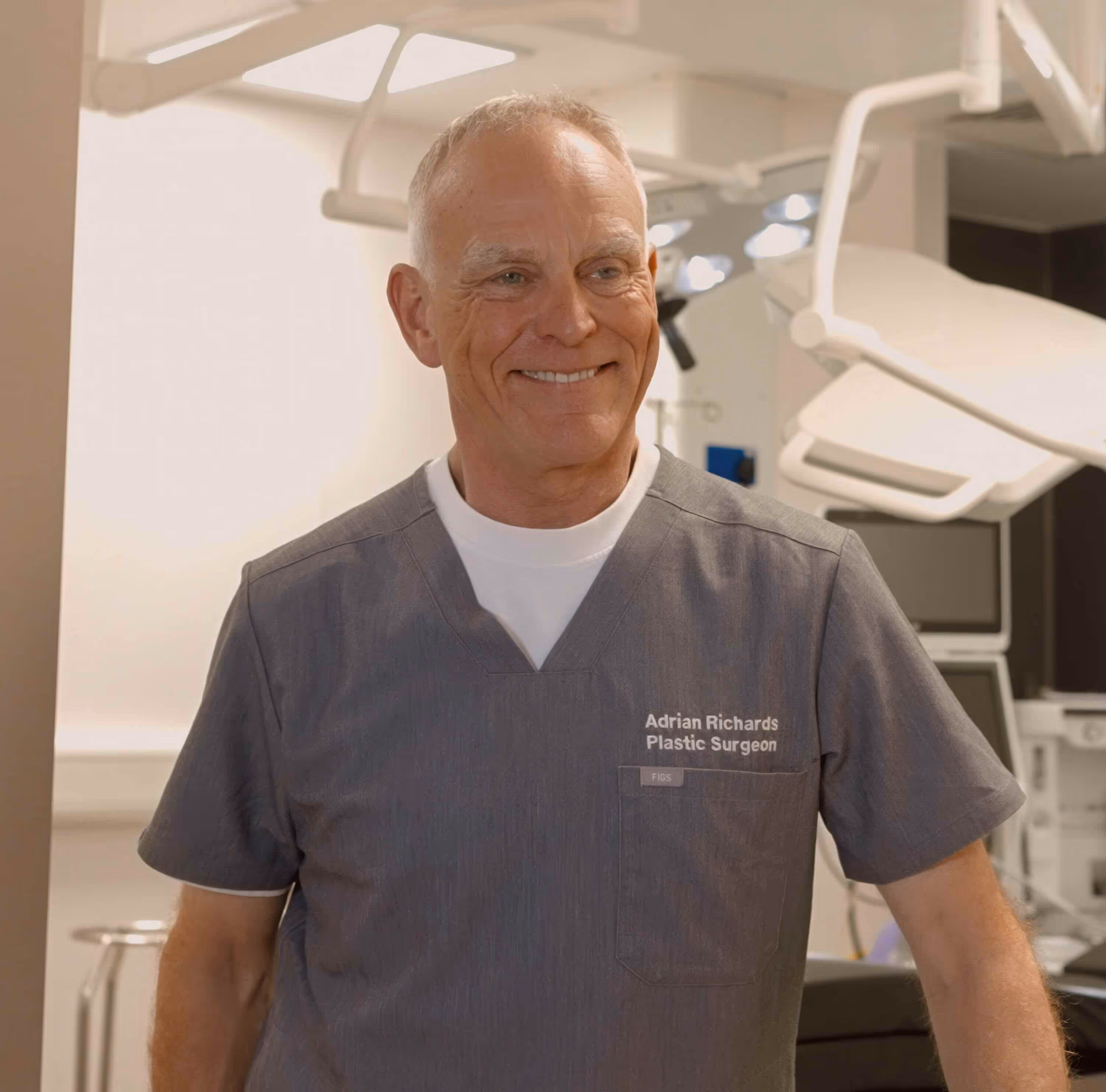 Smiling male plastic surgeon wearing gray scrubs in a medical office.