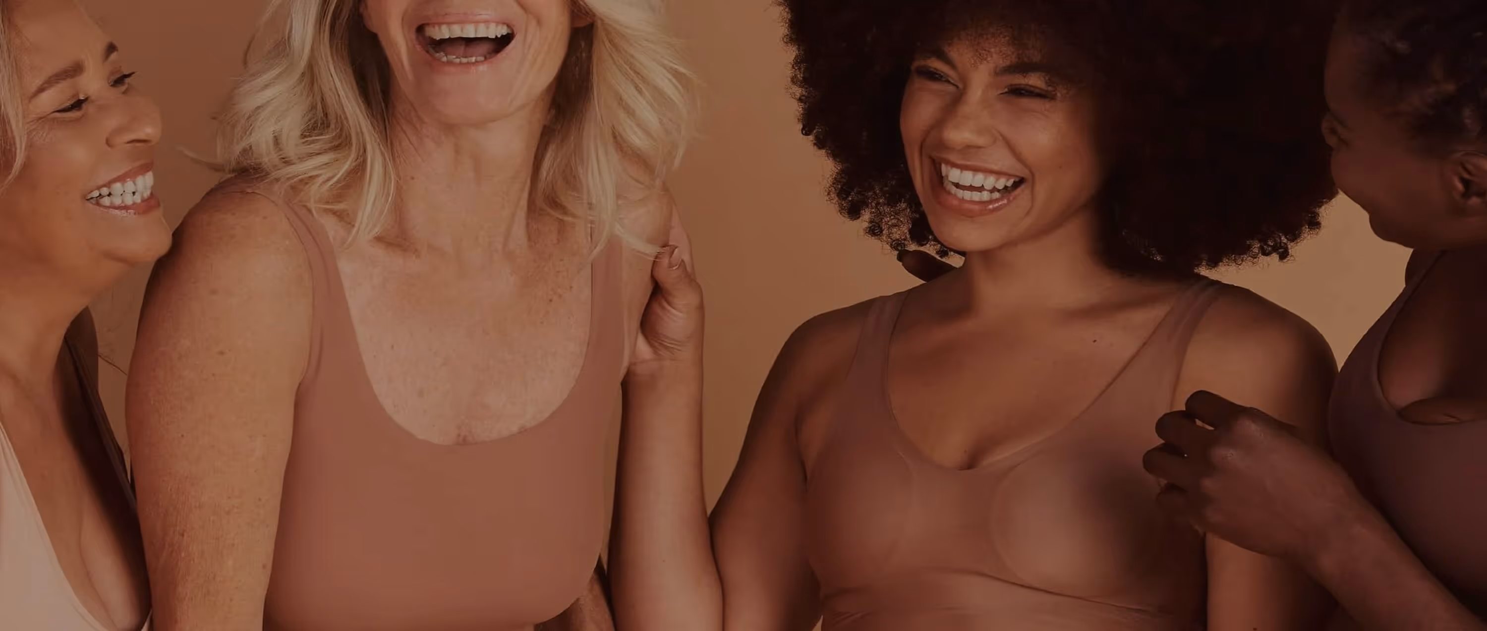 Four diverse women smiling and laughing together wearing beige tops against a neutral background.