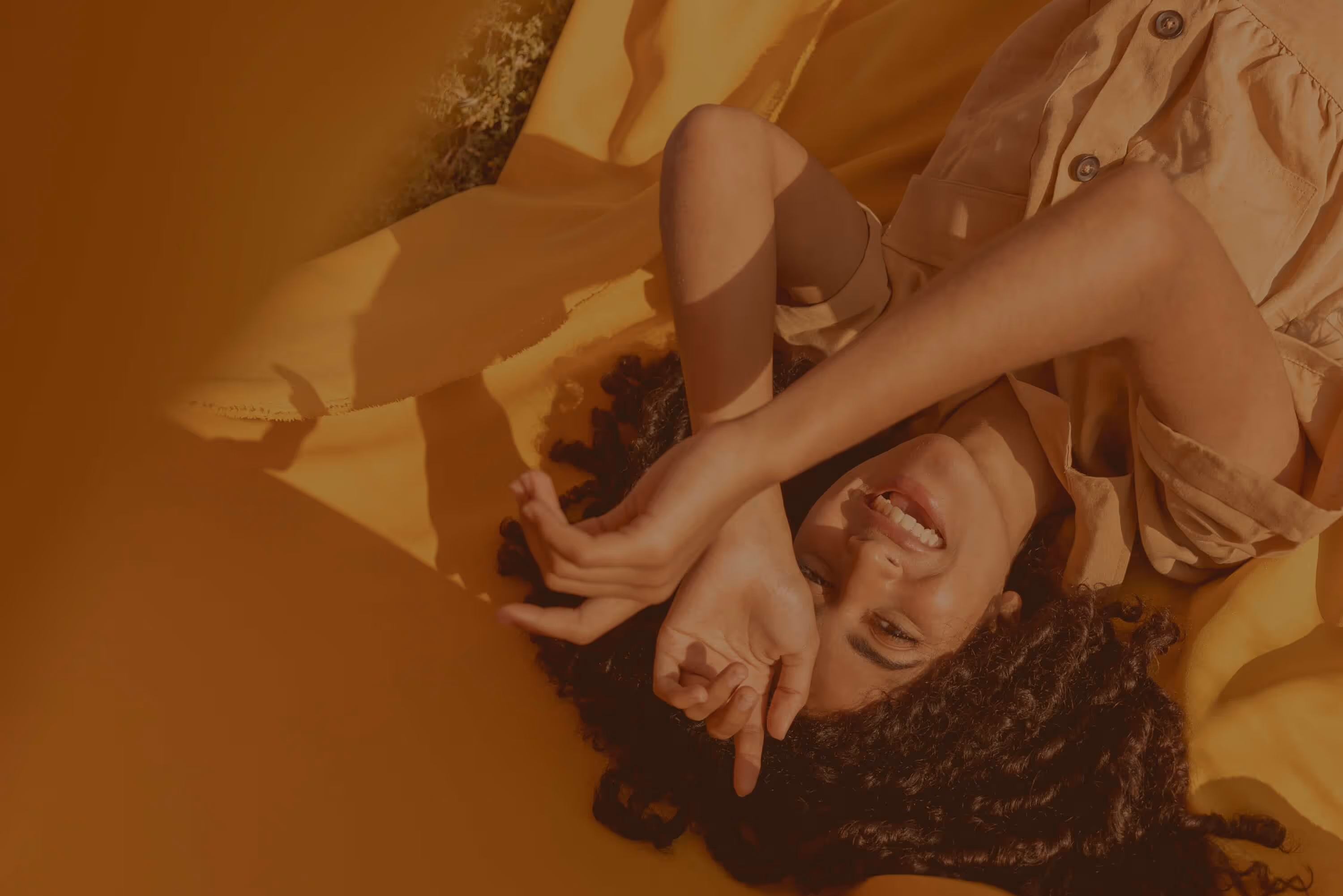 Smiling woman with curly hair lying on a yellow blanket with arms raised over her head.