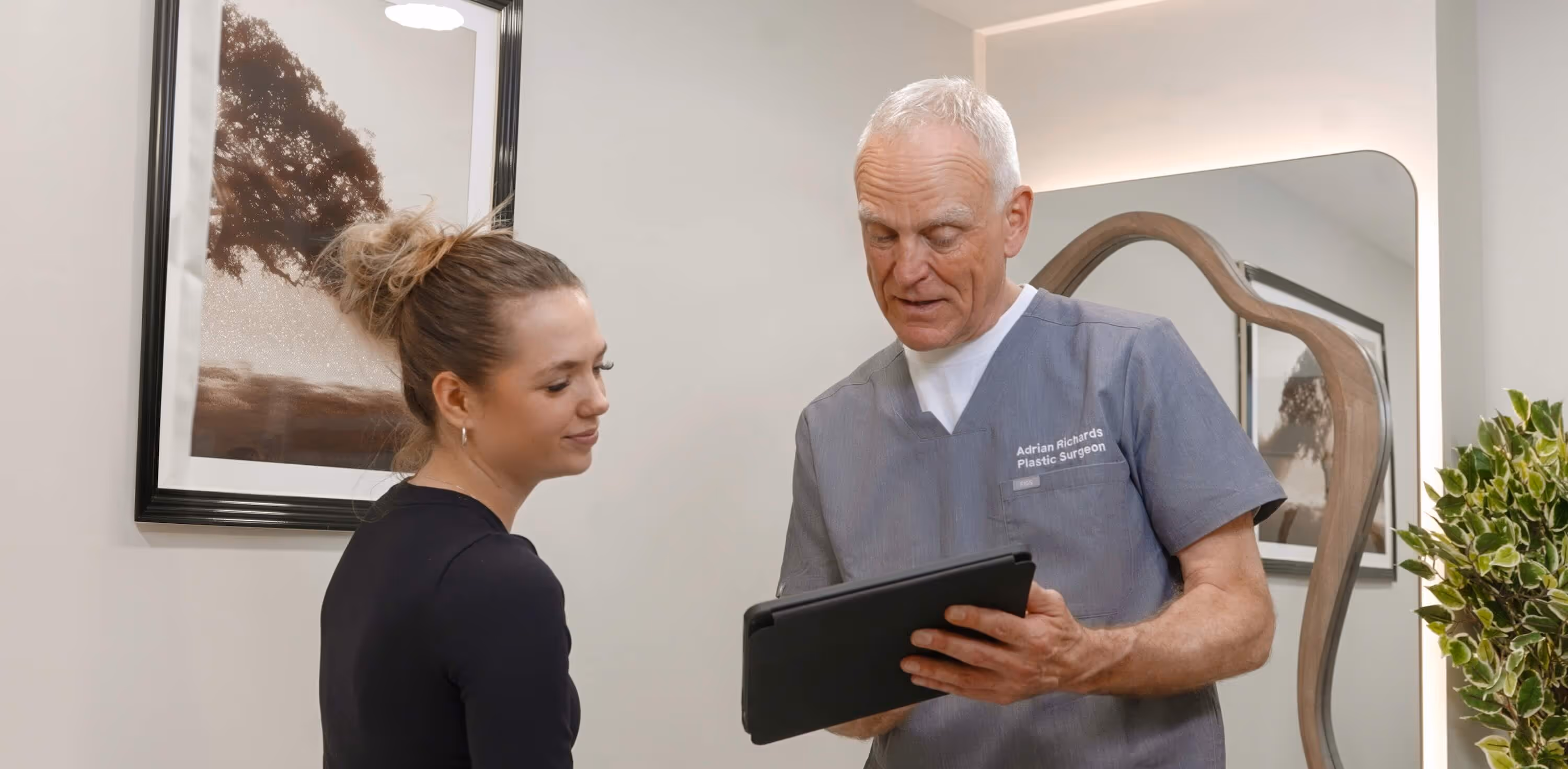 Plastic surgeon Adrian Richards shows a tablet to a smiling female patient in a consultation room.