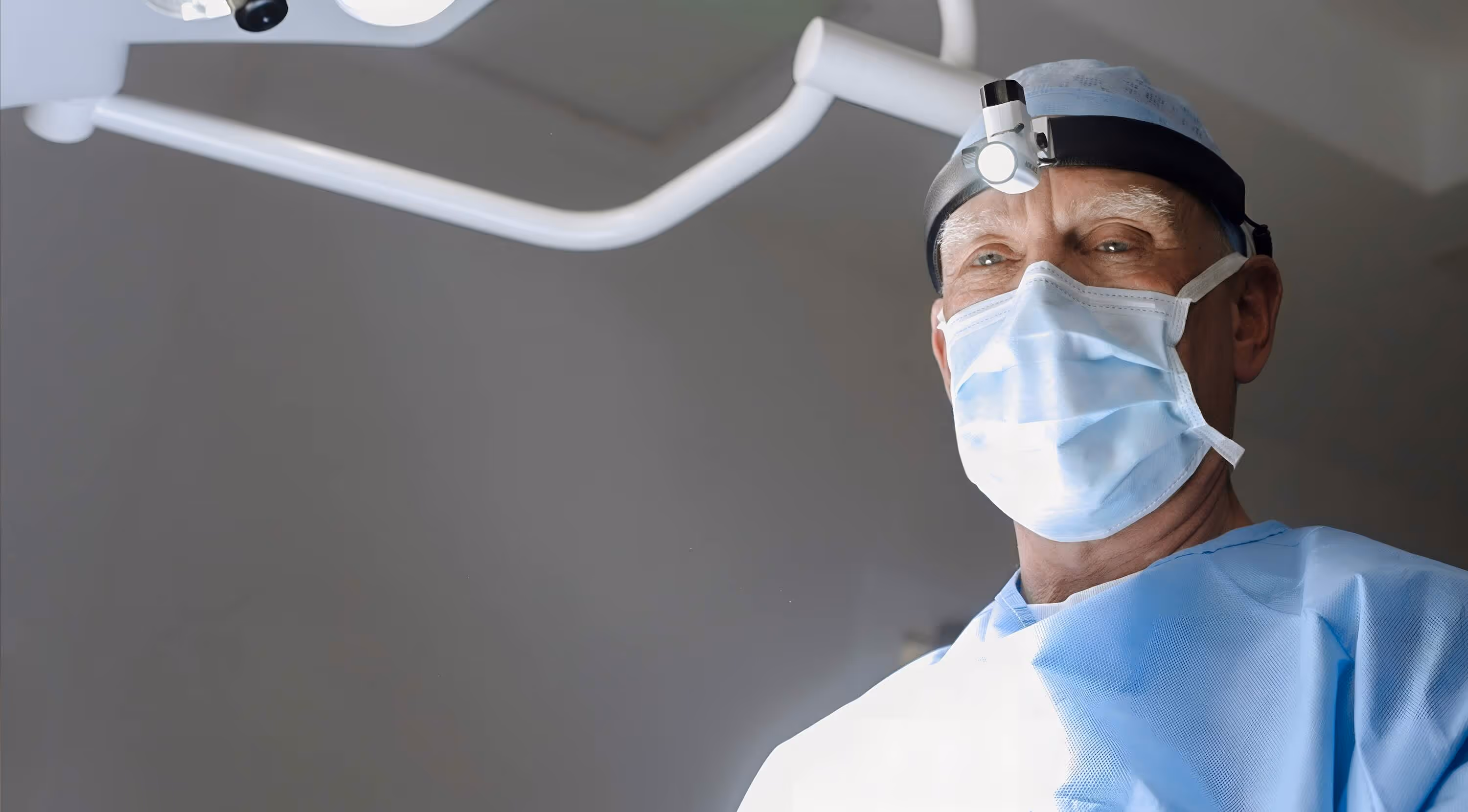 Male surgeon wearing surgical mask, cap, and headlight in an operating room.
