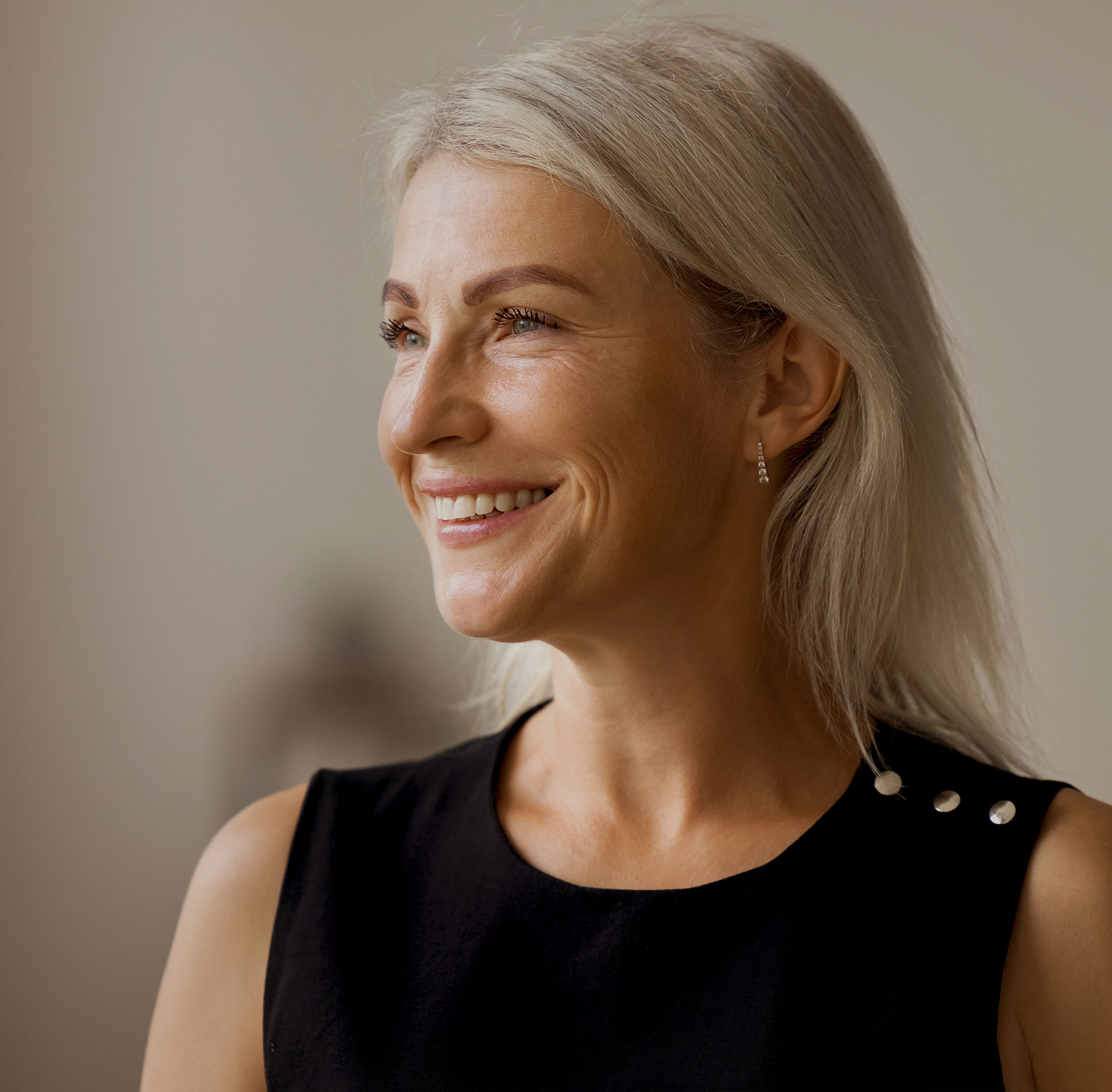 Smiling mature woman with long blonde hair wearing a black sleeveless top, looking to the side in a softly lit indoor setting.