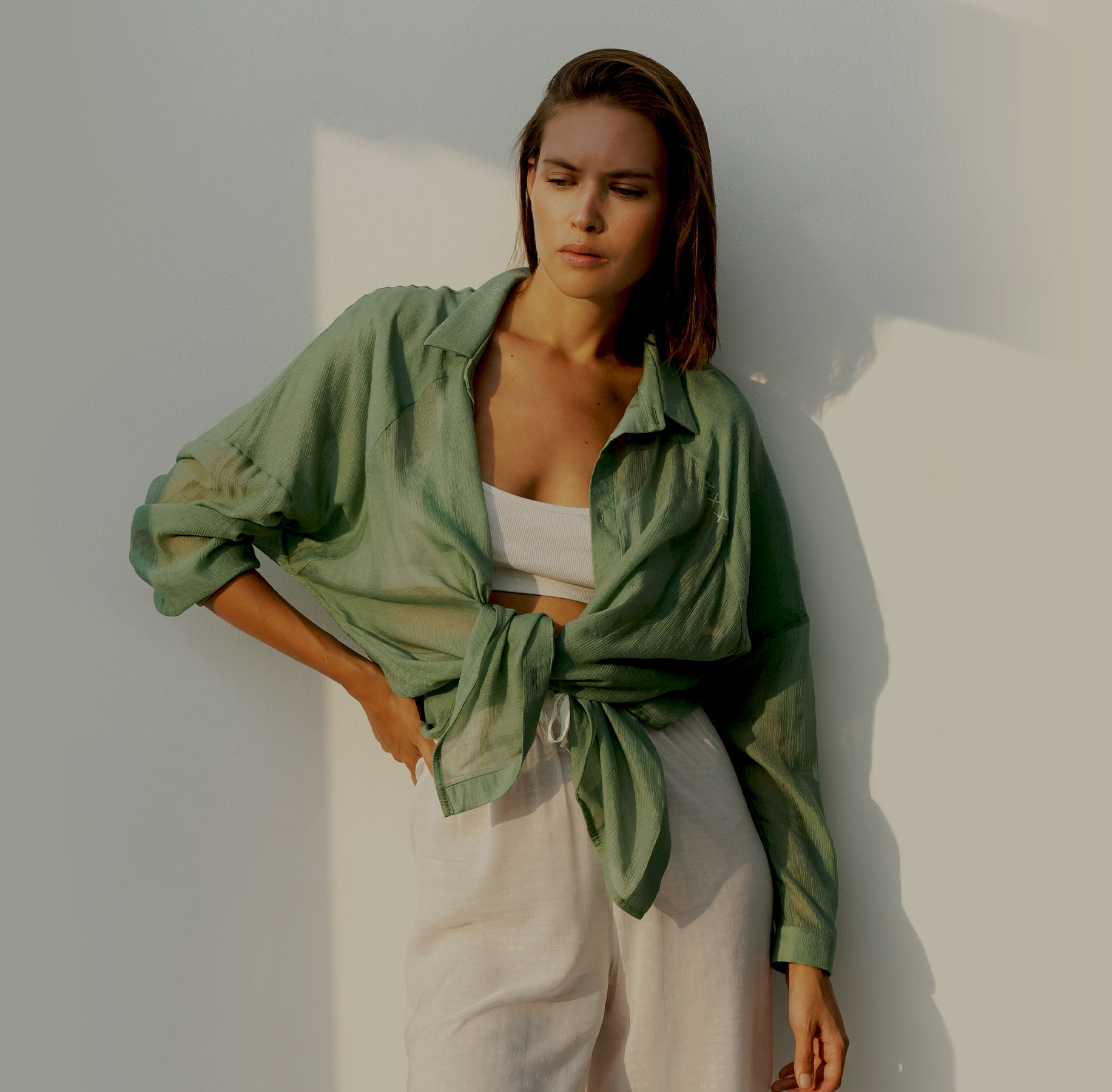 A woman in a loose green shirt and white pants poses confidently against a light wall.