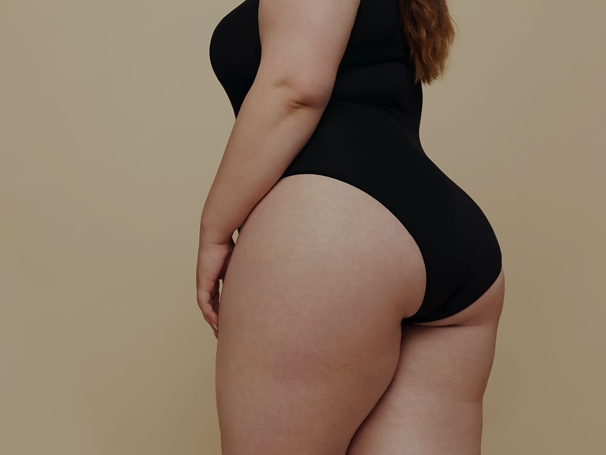 A woman in a black bodysuit poses sideways against a neutral brown background, showcasing her silhouette.