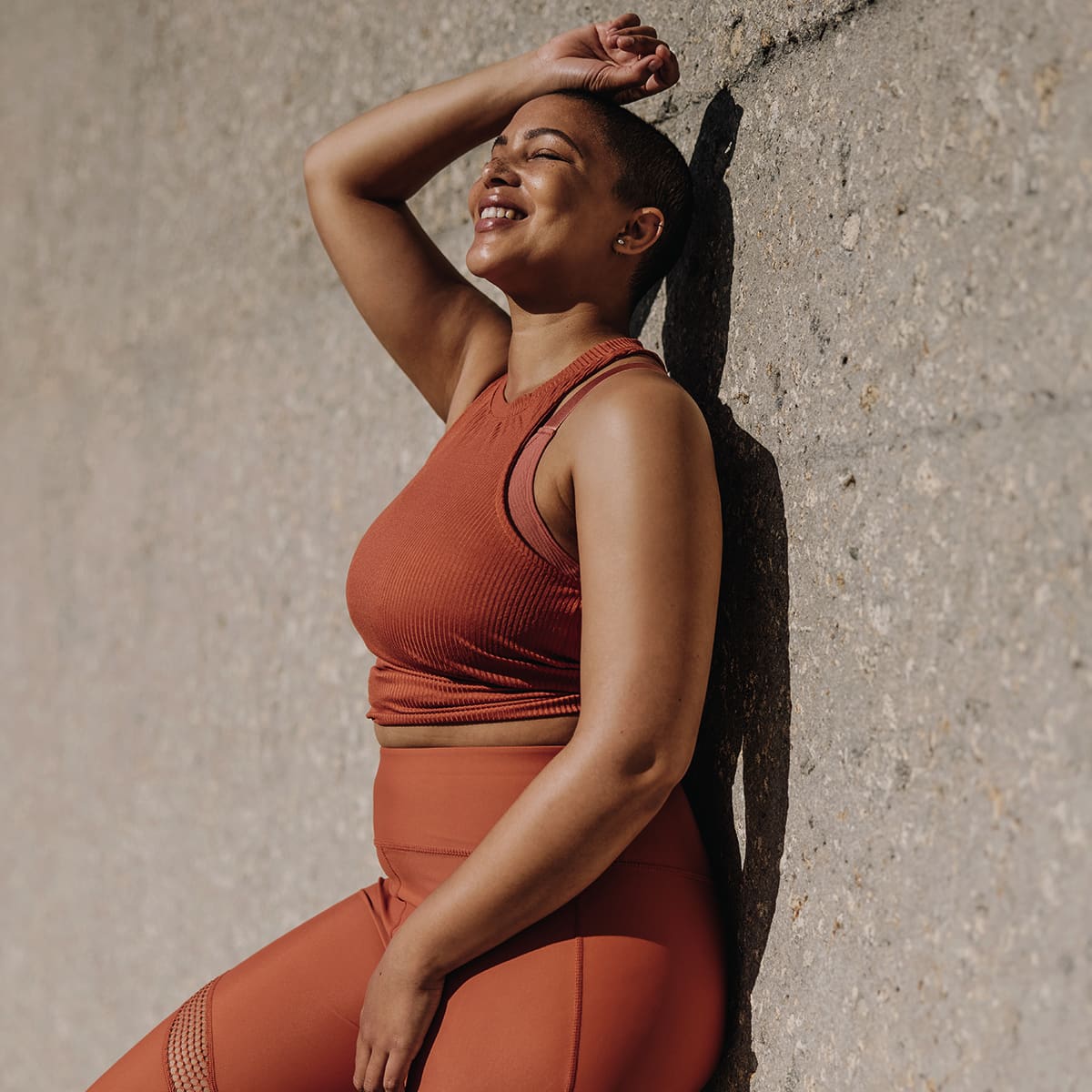 Smiling woman in a rust-colored athletic outfit sitting against a textured wall, showcasing a confident and carefree attitude.