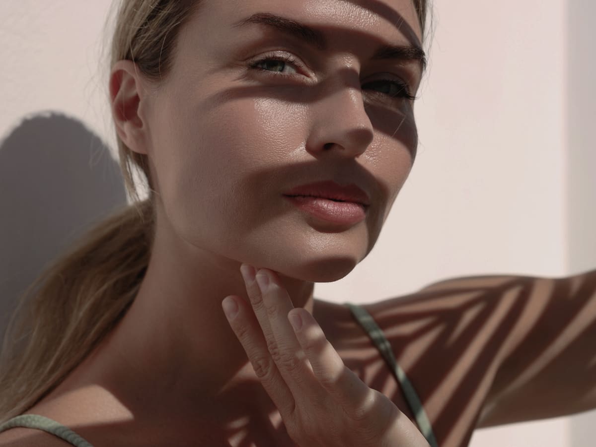 A woman with long blonde hair gently touches her chin while shadows from palm fronds fall across her face, accentuating her features.