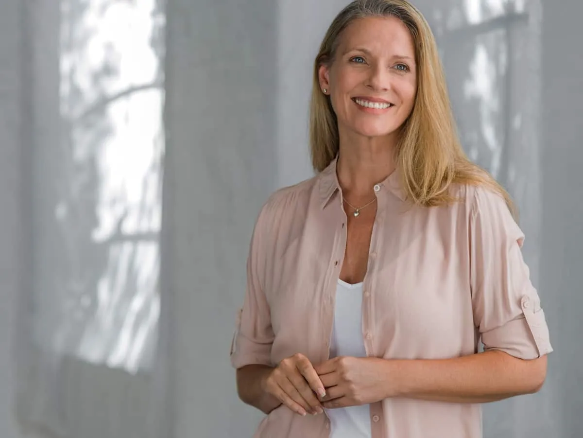 Smiling woman with long blonde hair wearing a light pink blouse, standing in front of a softly lit background.