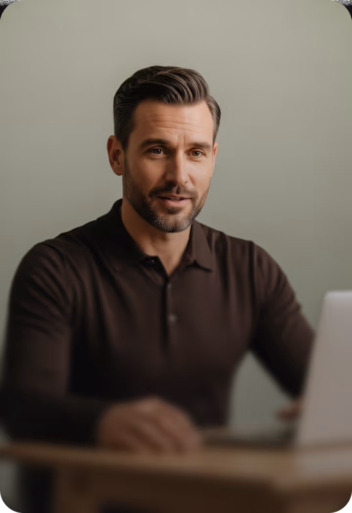 Man with styled dark hair and beard wearing a dark shirt, looking at a laptop on a wooden table.