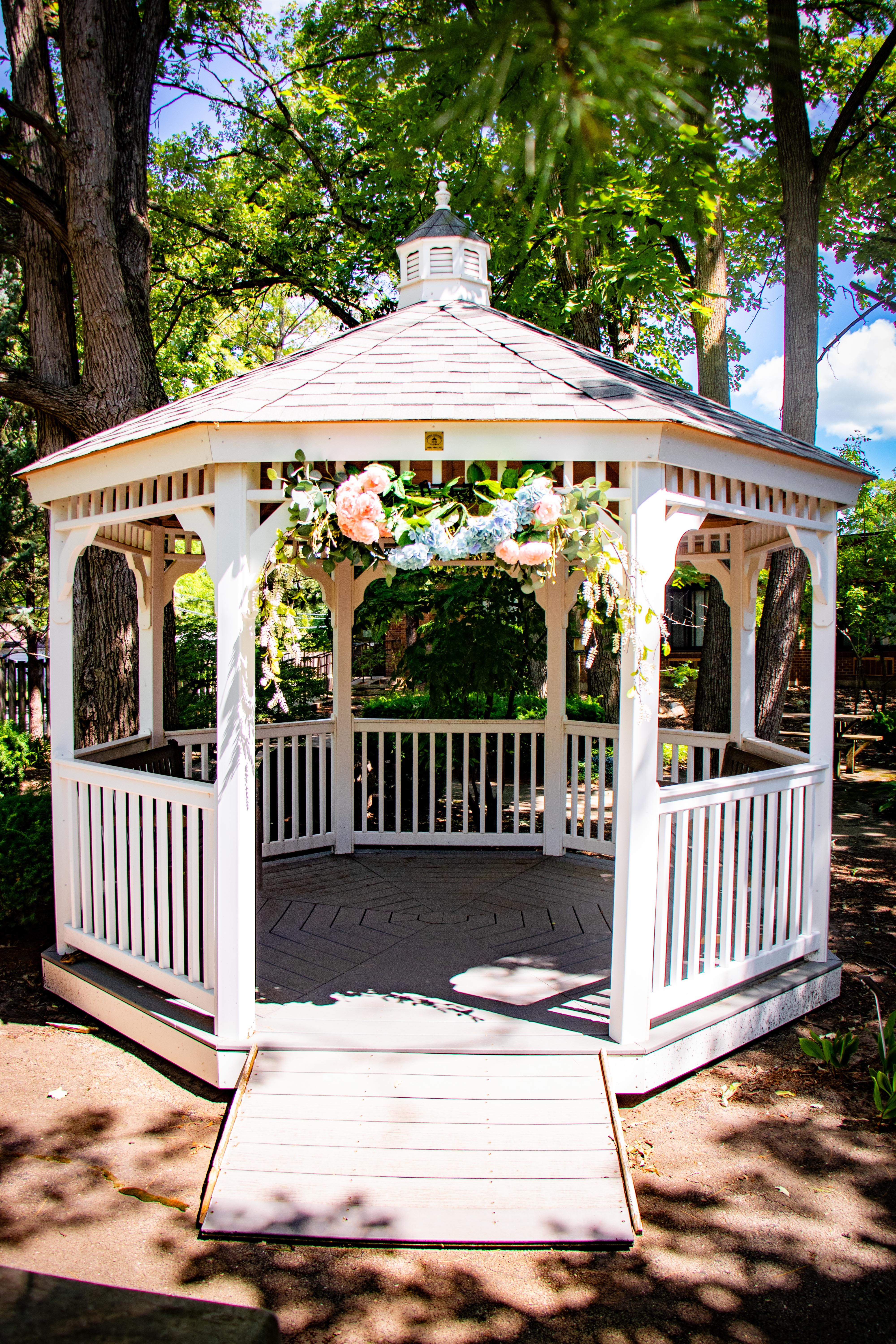 White wooden gazebo with a floral wreath decoration in a sunlit green park setting.